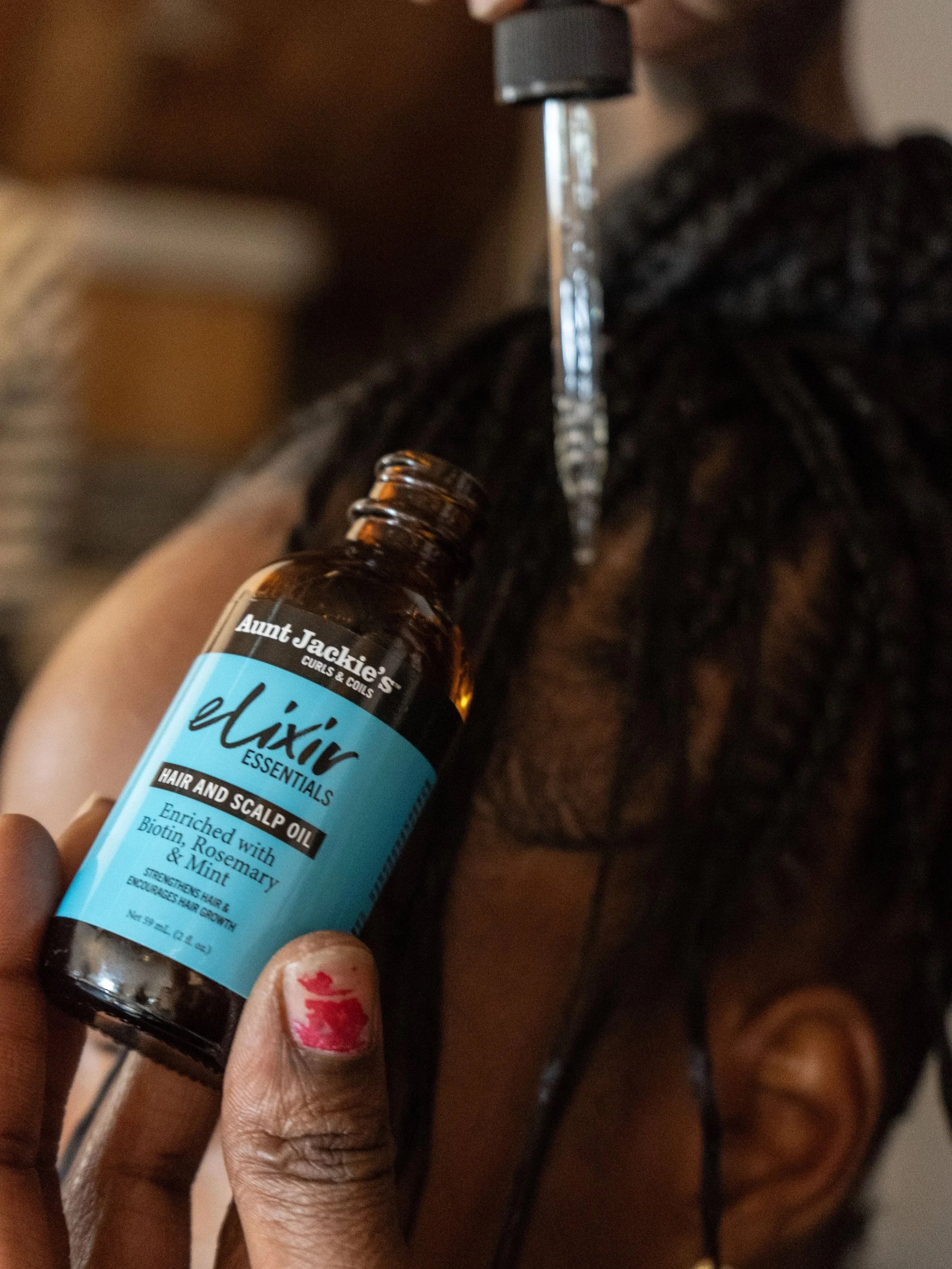 A person using a scalp oil on their dreadlocks with a hair dyeing tool nearby, holding a bottle of Aunt Jackie's Daxir Essentials Hair and Scalp Oil enriched with biotin, rosemary, and mint.