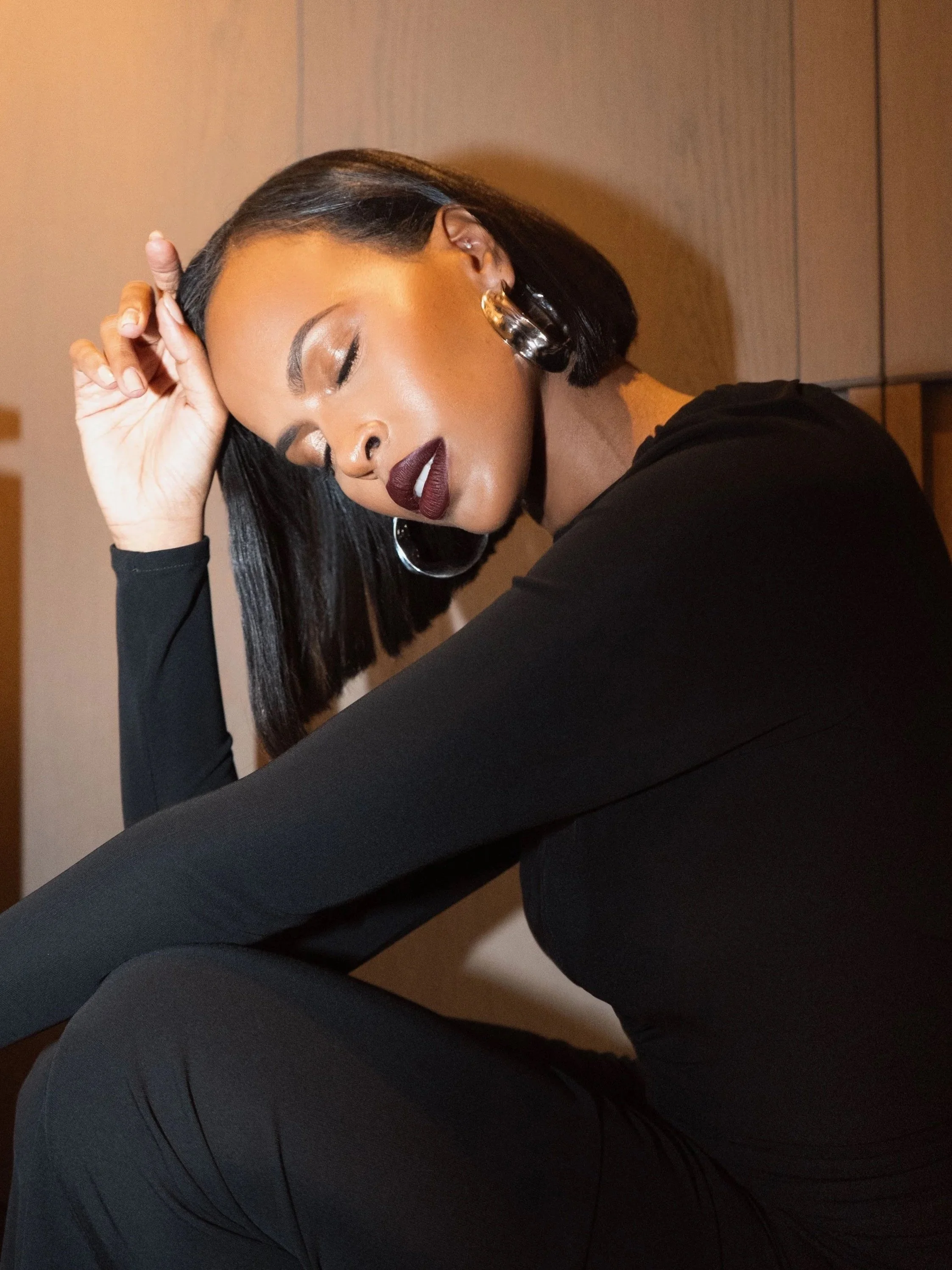 A woman with dark hair, closed eyes, and dark lipstick, resting her head on her hand, wearing hoop earrings and a black long-sleeve top, seated against a wall.