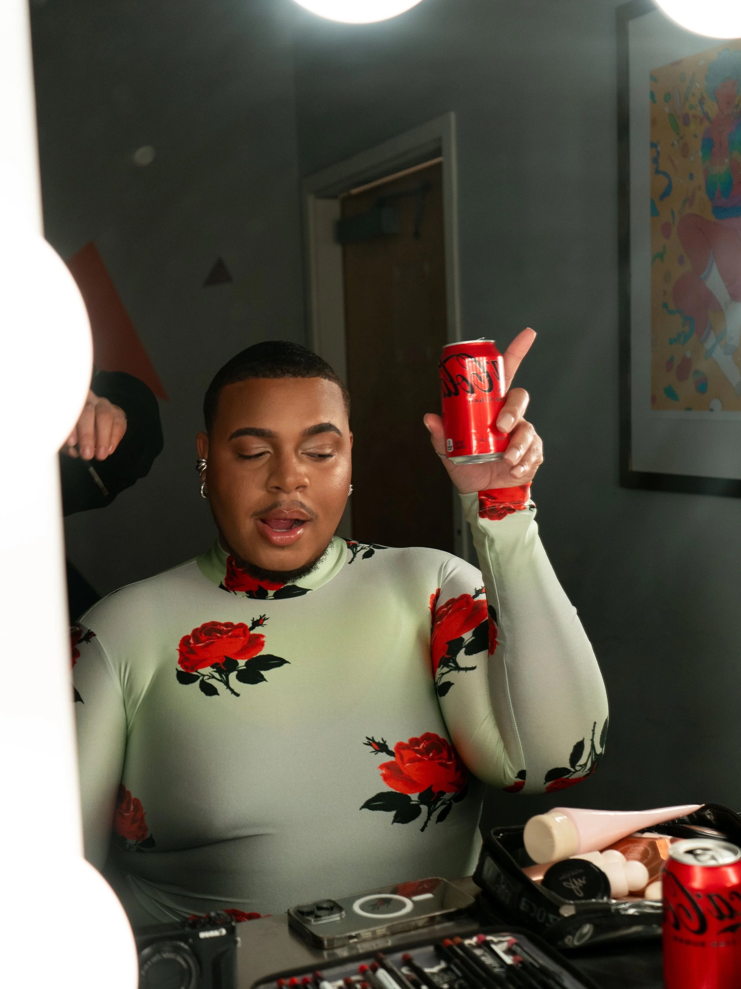 A person with short hair and earrings, wearing a light green top with red flowers, holding a red soda can and pointing upward with their index finger, standing in front of a mirror surrounded by makeup and beauty products.