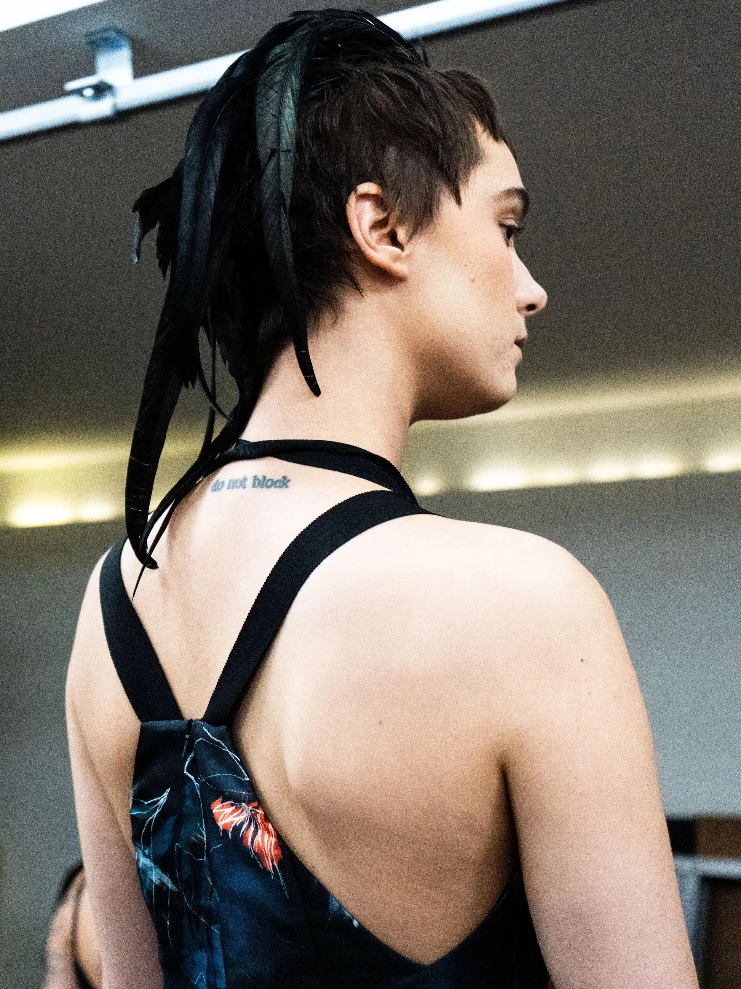 A young woman with short dark hair and feathers in her hair, wearing a black dress with floral details and a strap around her neck, seen from the side in an indoor setting.