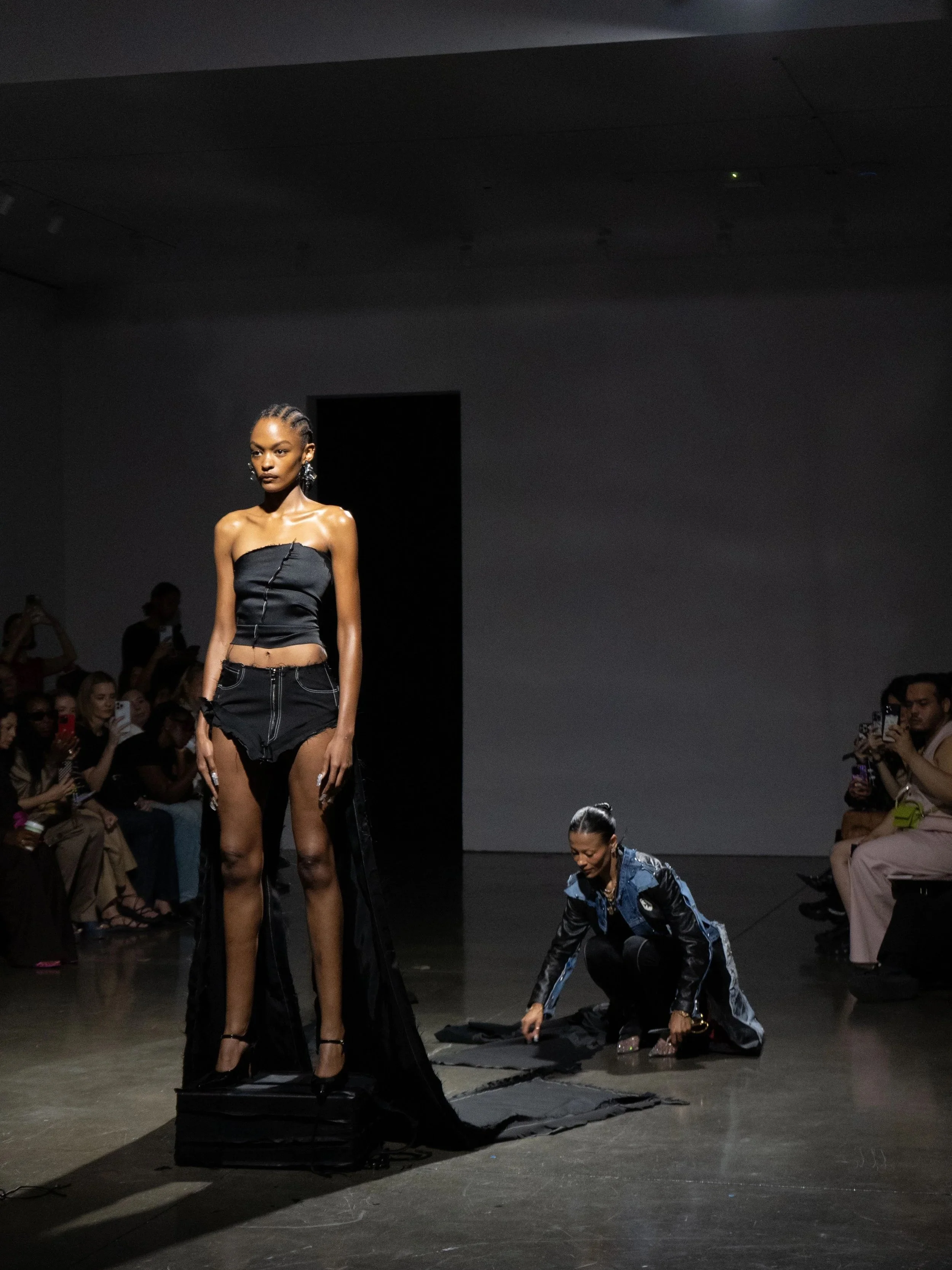 A fashion runway show with a dark-skinned female model standing on a platform, wearing a black strapless top and black high-slit shorts, with an assistant kneeling beside her adjusting fabric on the runway.