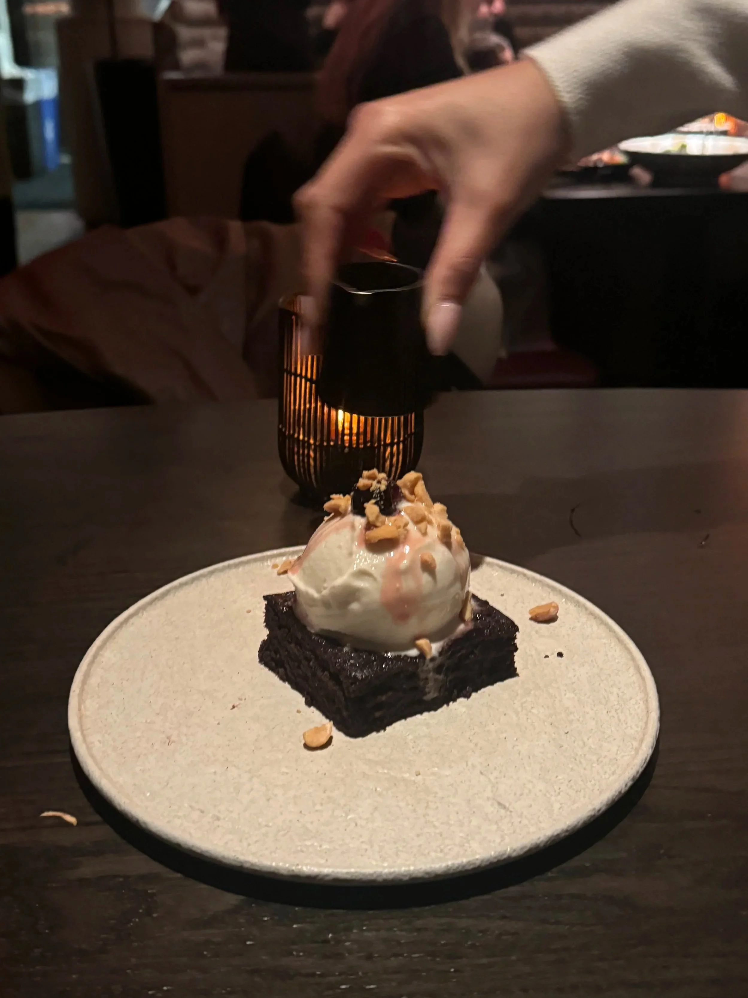 A dessert with a brownie base topped with a scoop of vanilla ice cream, garnished with nuts and a cherry, on a round beige plate. A hand is pouring a sauce from a small container over the ice cream. In the background, there is a candle holder on a dark table in a dimly lit restaurant.