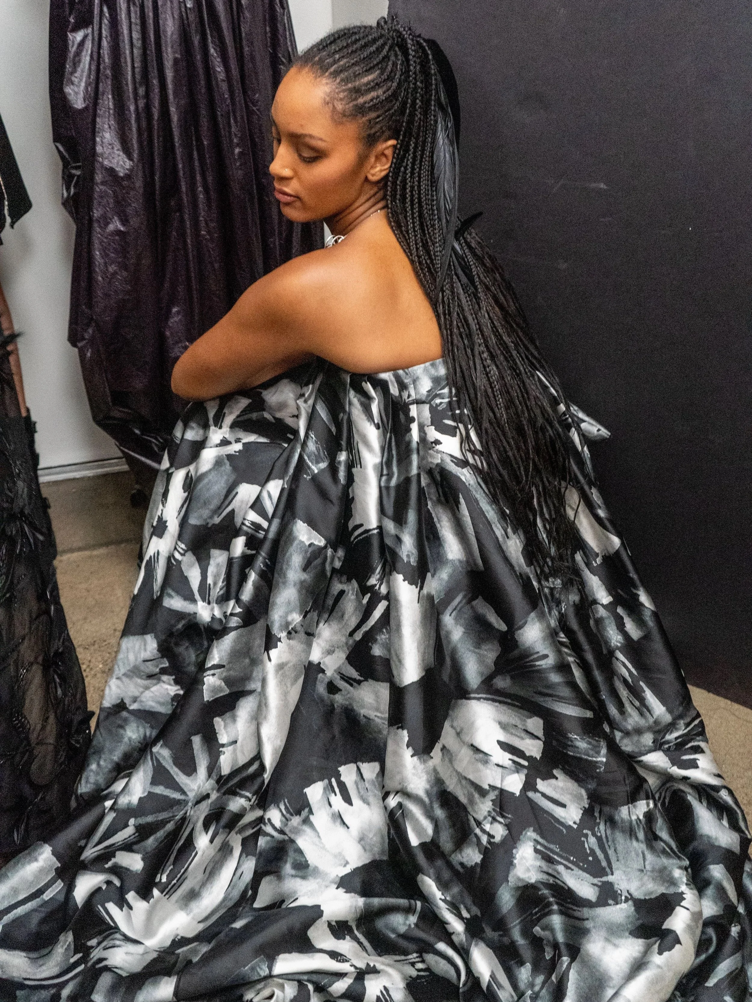 A woman with long braided hair sitting on the floor, wearing a black and white patterned dress, with her arms crossed over her knees, leaning against a black wall.