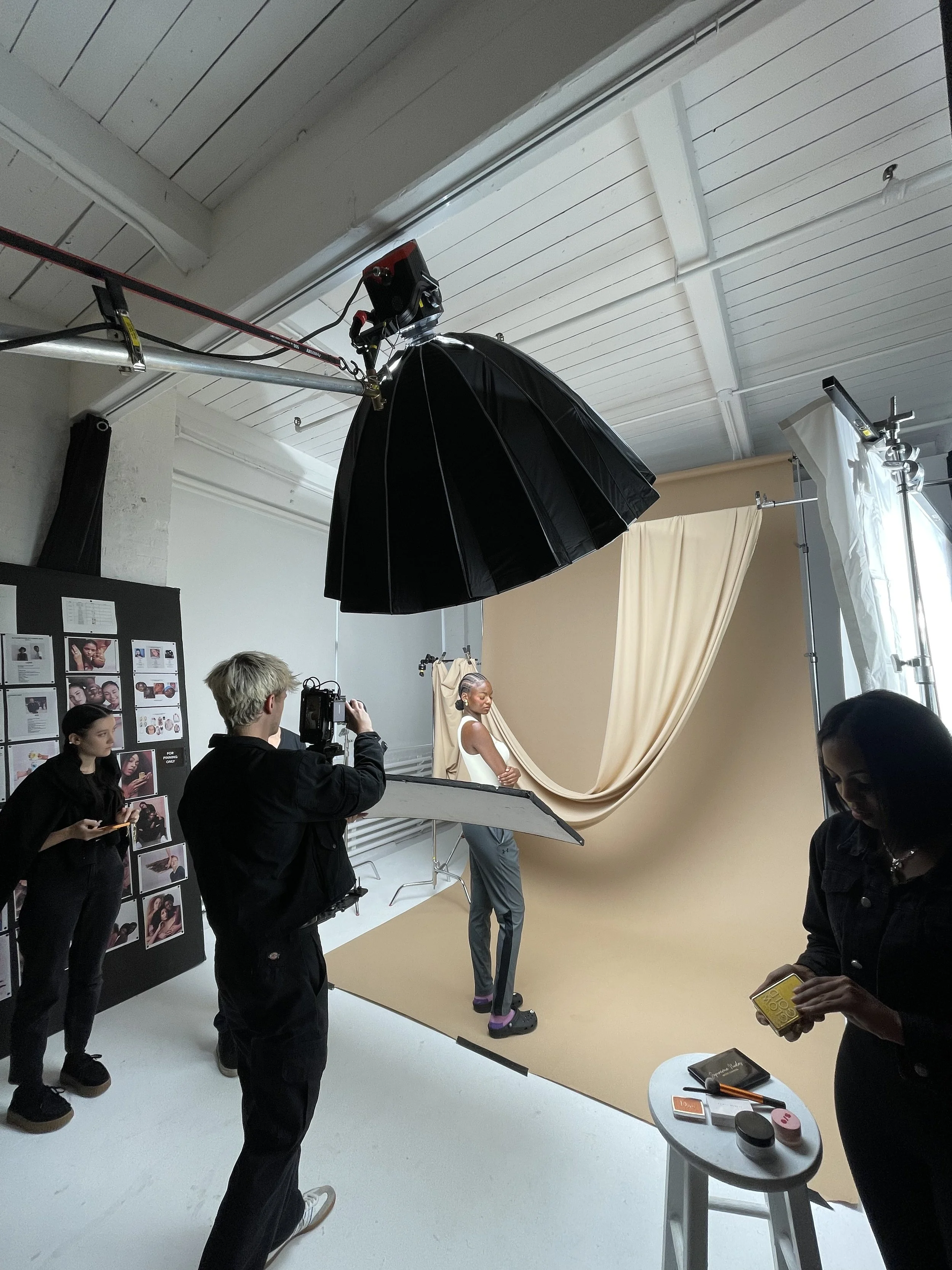 A photoshoot studio with a model posing against a beige backdrop, surrounded by crew members and equipment, including a large black umbrella light, a camera, and reflectors.