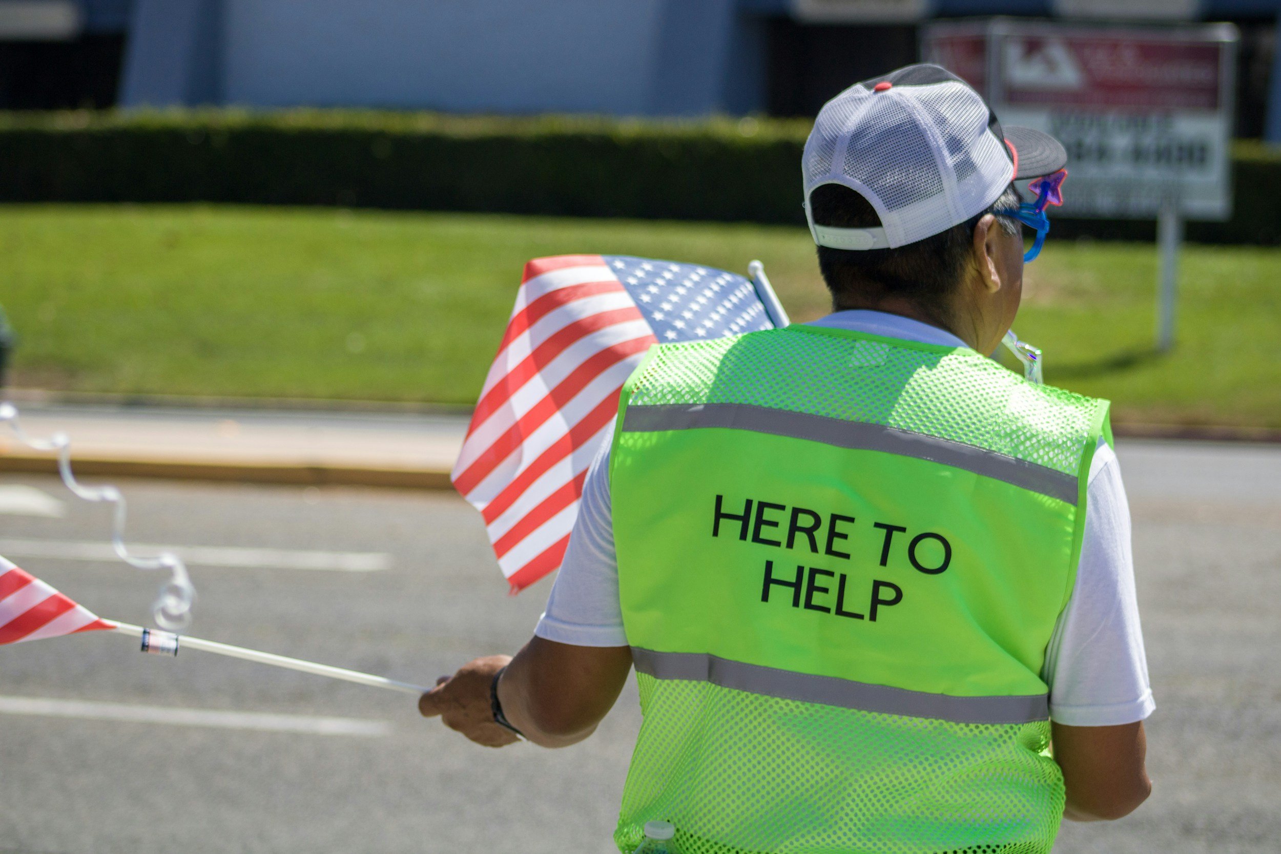A person wearing a neon green safety vest with the text "HERE TO HELP" on the back, a gray and black baseball cap, and star-shaped sunglasses, standing outdoors near a road with a grassy lawn in the background.
