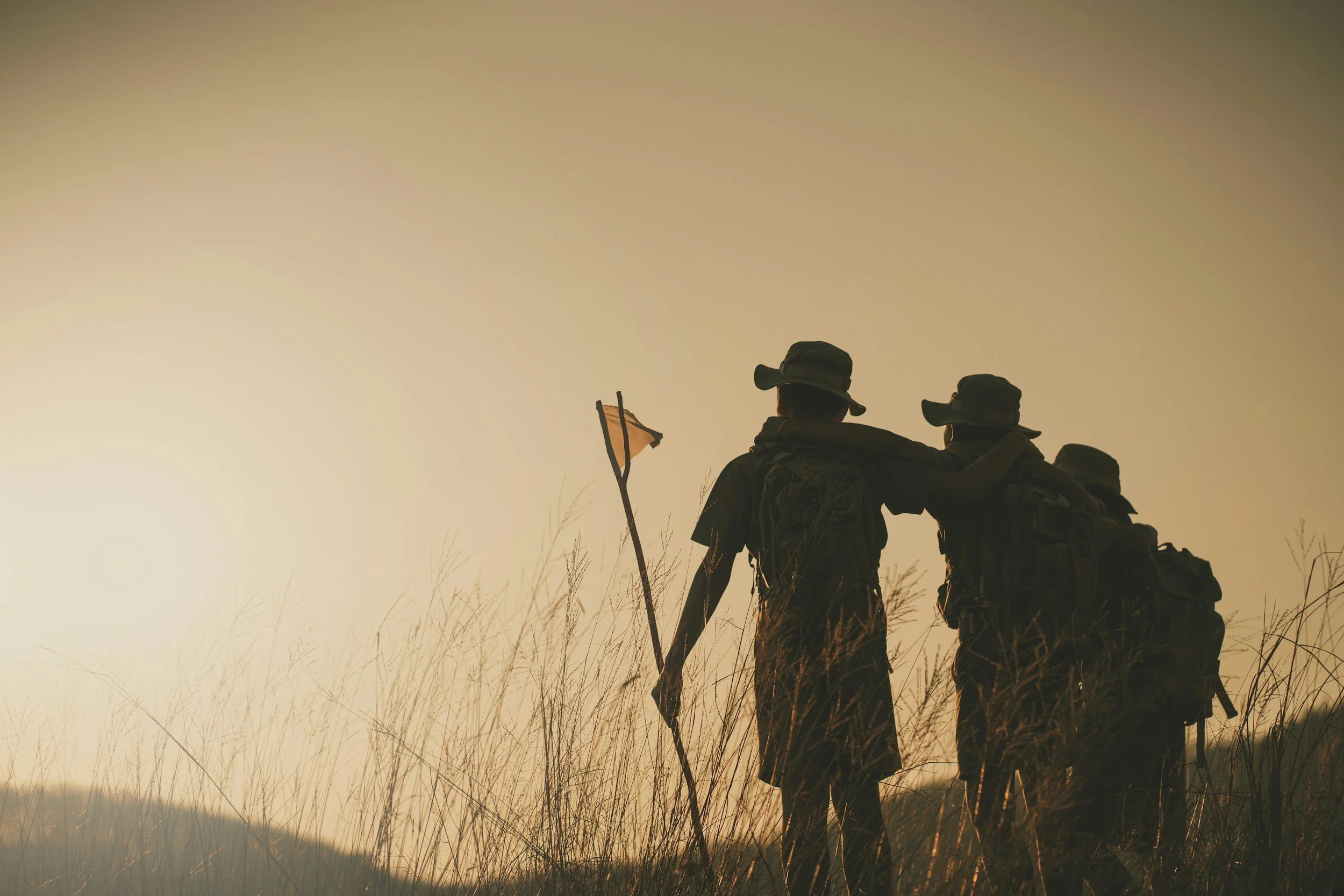 Two hikers with backpacks and hats standing together on a grassy hill at sunset, one of them holding a walking stick, looking out at the view.