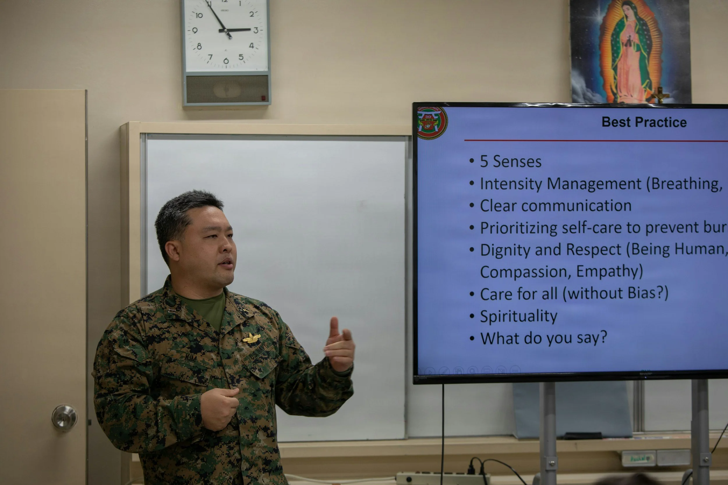 A man in military uniform giving a presentation in front of a screen with a PowerPoint slide. The slide contains bullet points about best practices for communication and self-care.
