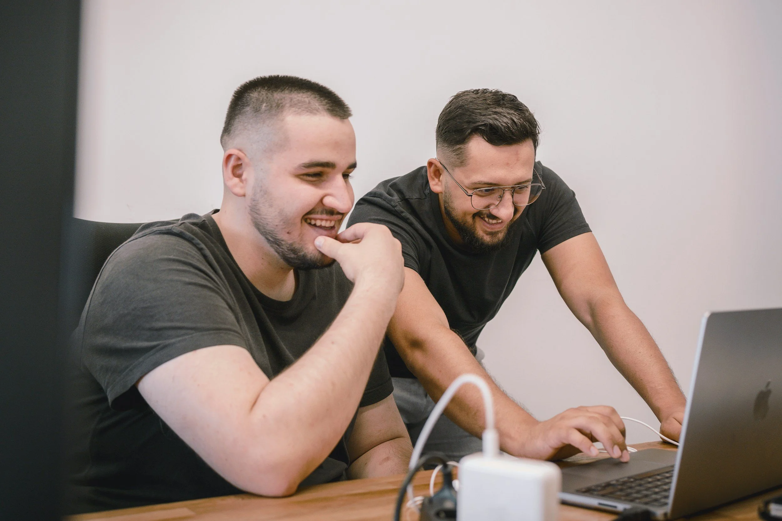 Two men sitting at a table, smiling, looking at a laptop, with charging cables and a power strip on the table.