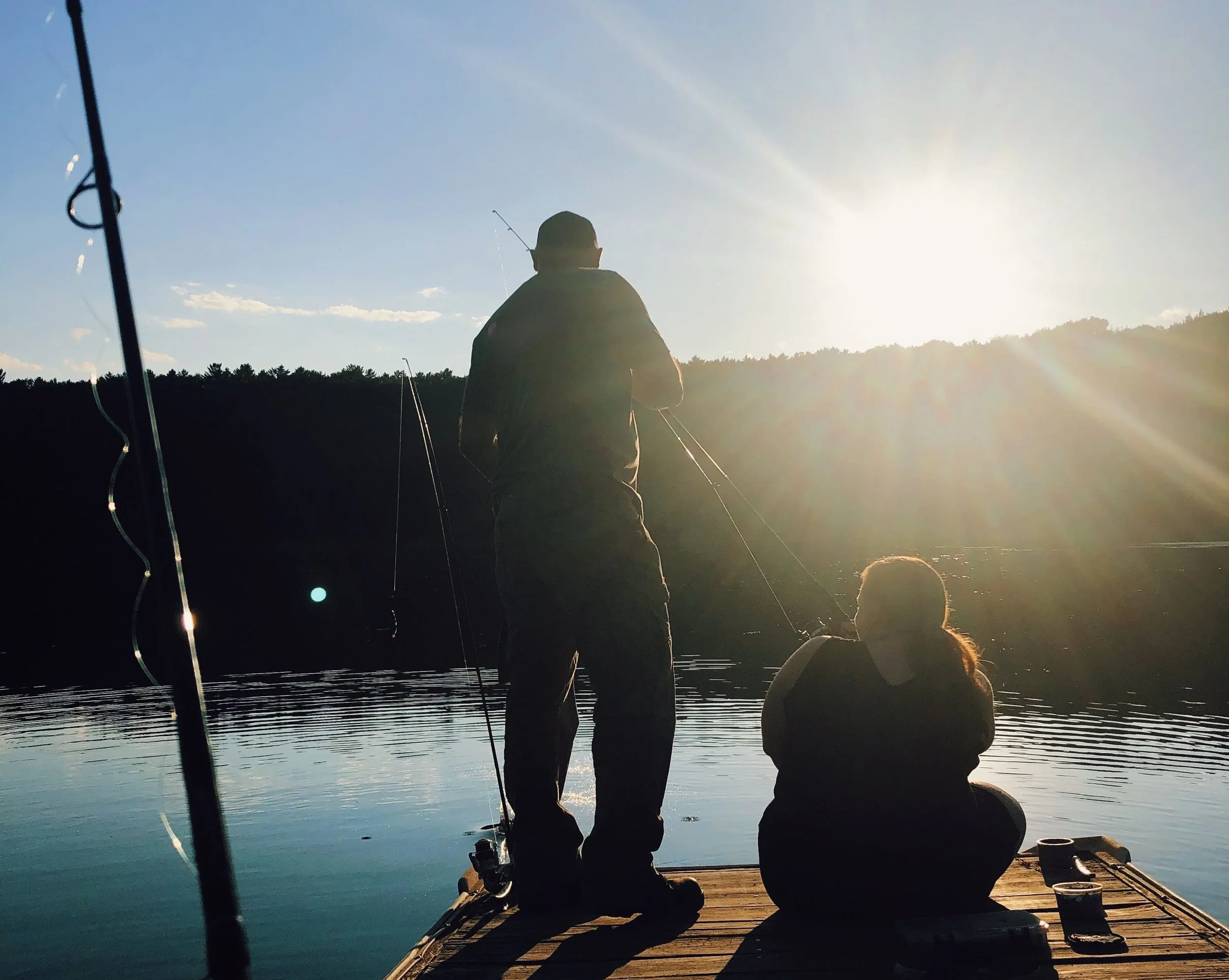 Two people fishing by a lake during sunset, one standing and one sitting with fishing rods set up nearby.