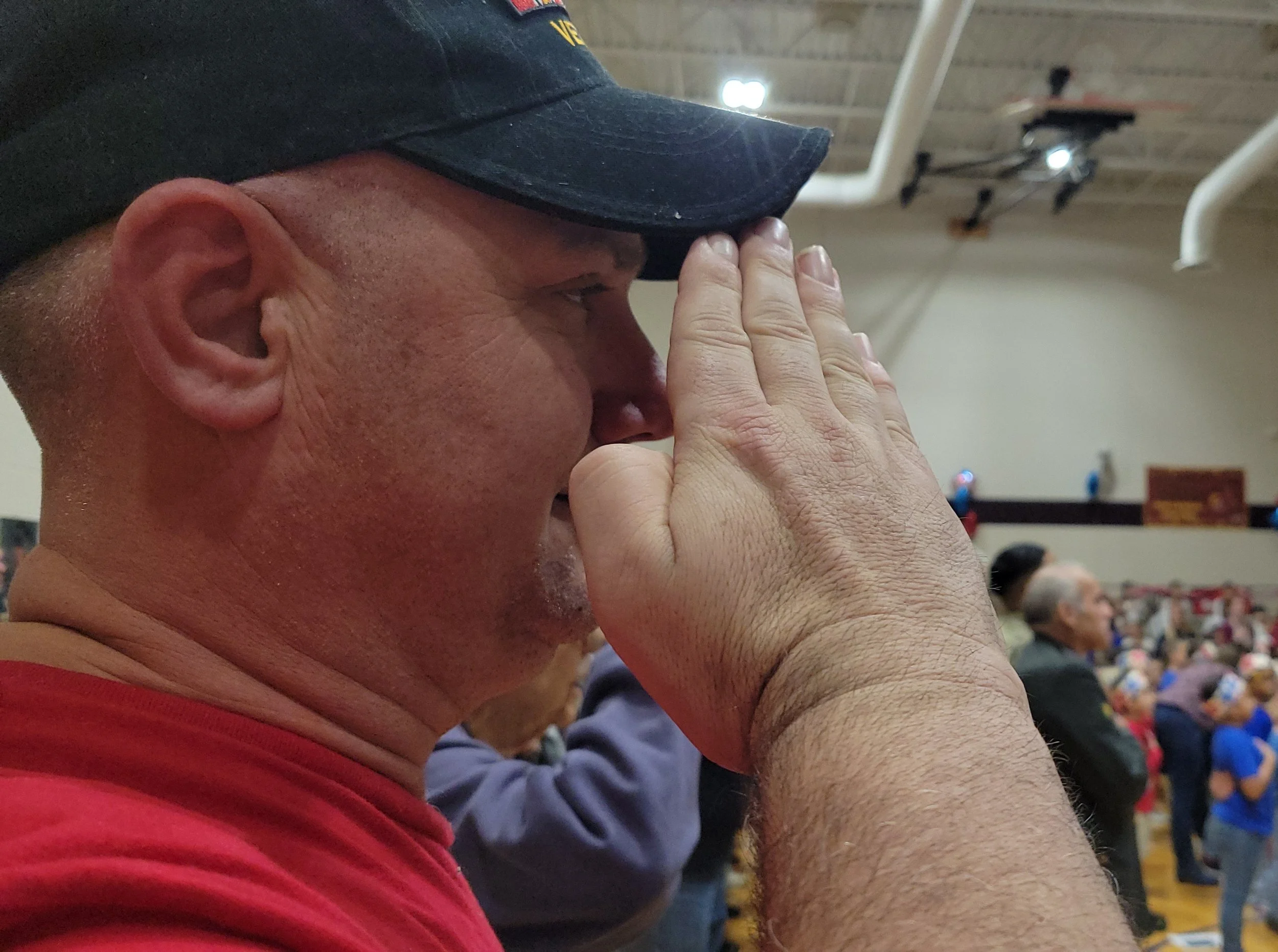 A man in a red shirt with a navy cap saluting, inside a gymnasium filled with people.