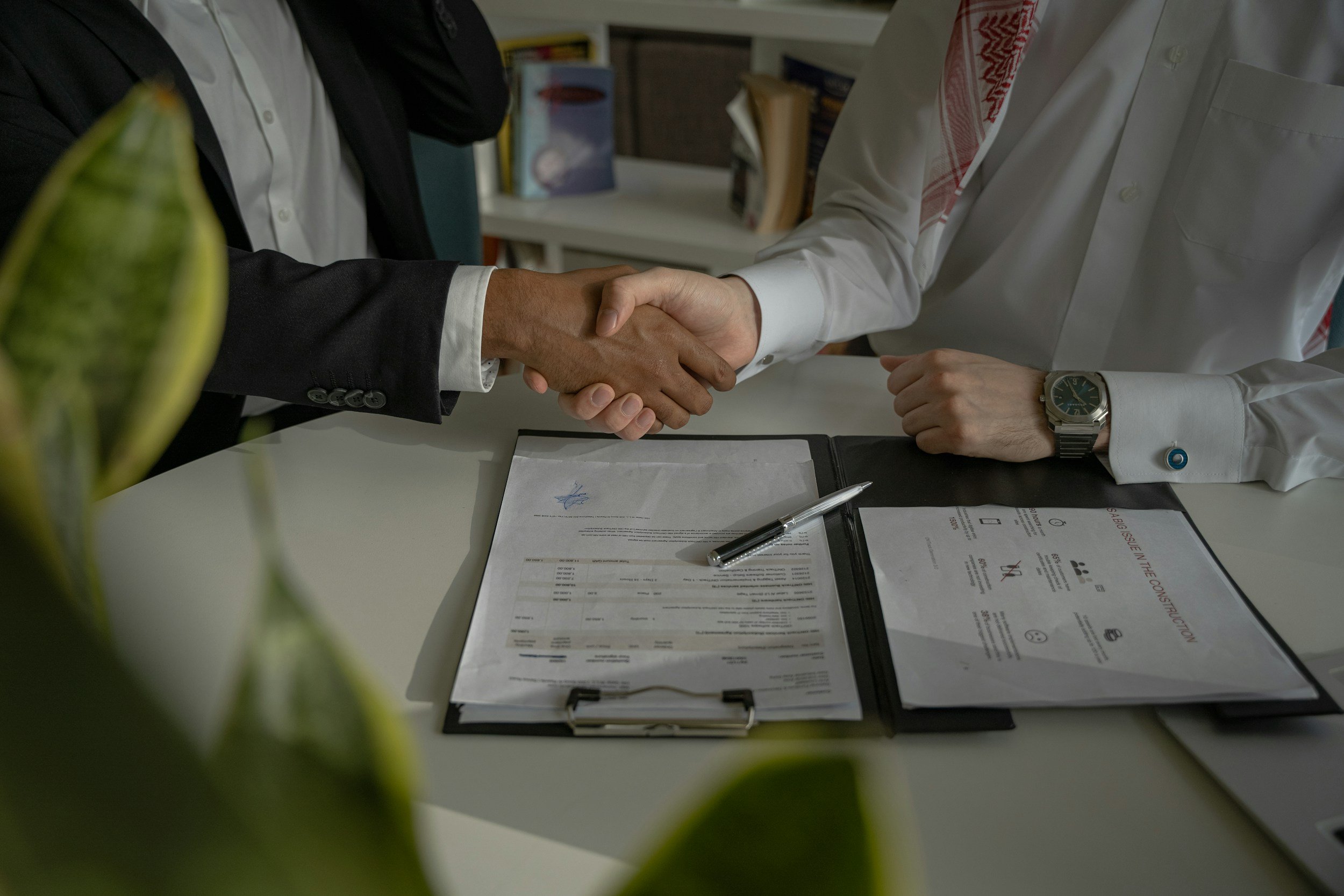 Two people shaking hands over a desk in an office, with documents, a pen, and a clipboard on the table. Plants are in the foreground.