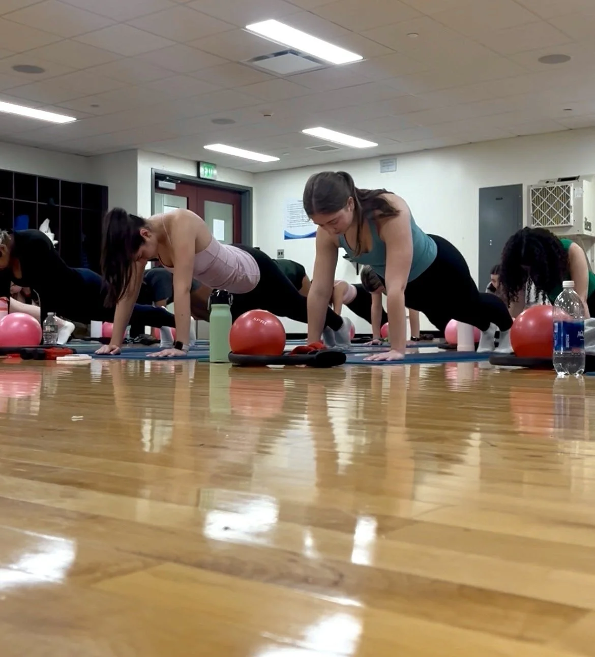 A group of women participating in a fitness class doing plank exercises on yoga mats in a gym, with water bottles and exercise balls beside them.
