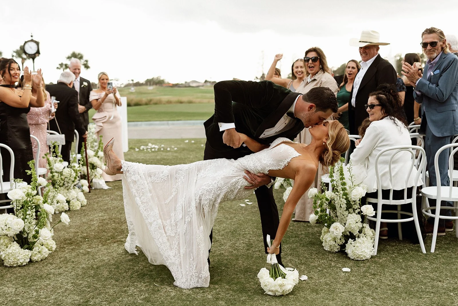 A newlywed couple shares a kiss at their outdoor wedding ceremony, with guests applauding and smiling around them amid floral decorations and a scenic background.