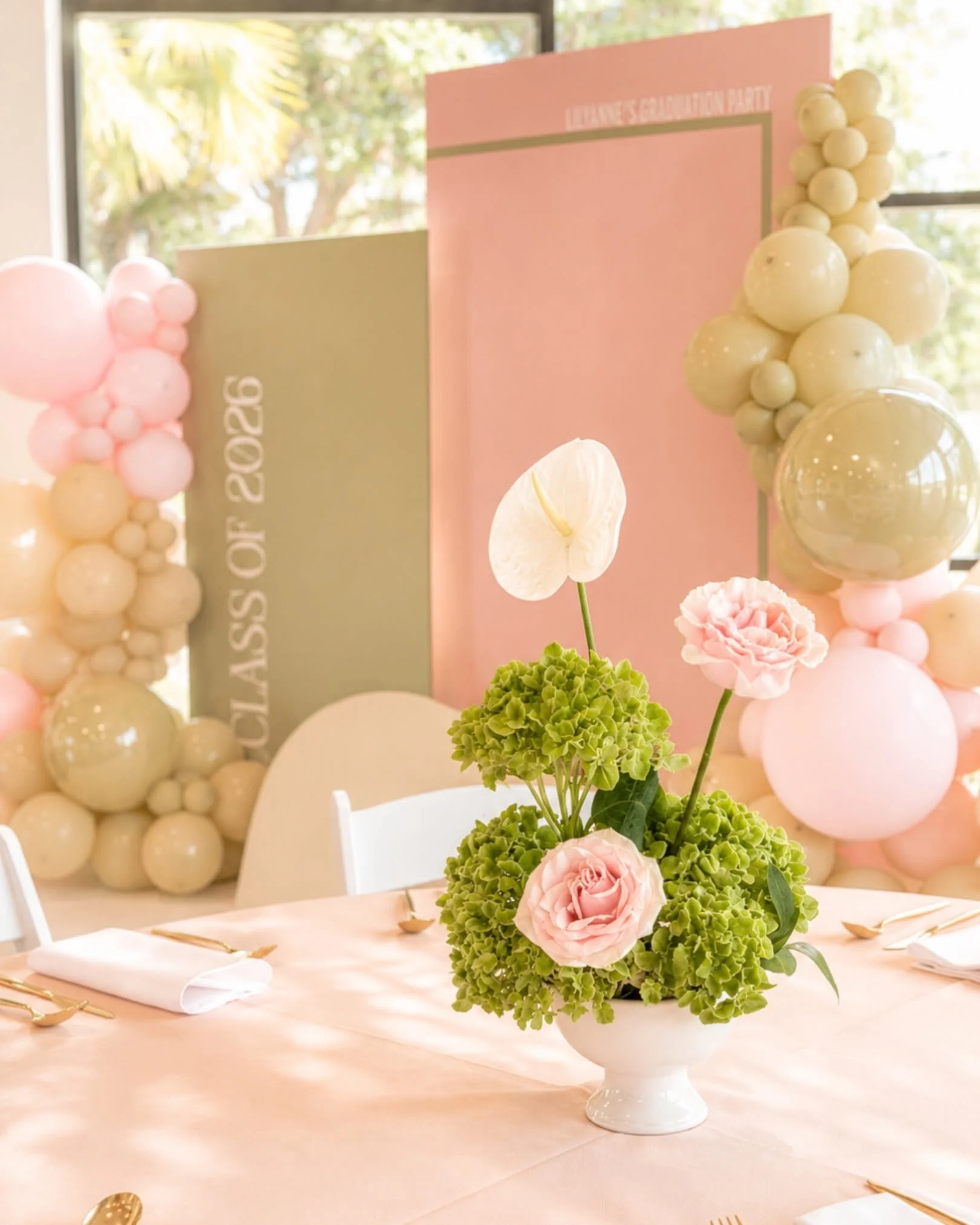 A table centerpiece featuring pink roses, green hydrangeas, and white anthuriums in a white bowl, set at a decorated event with pastel-colored balloons and signs in the background.
