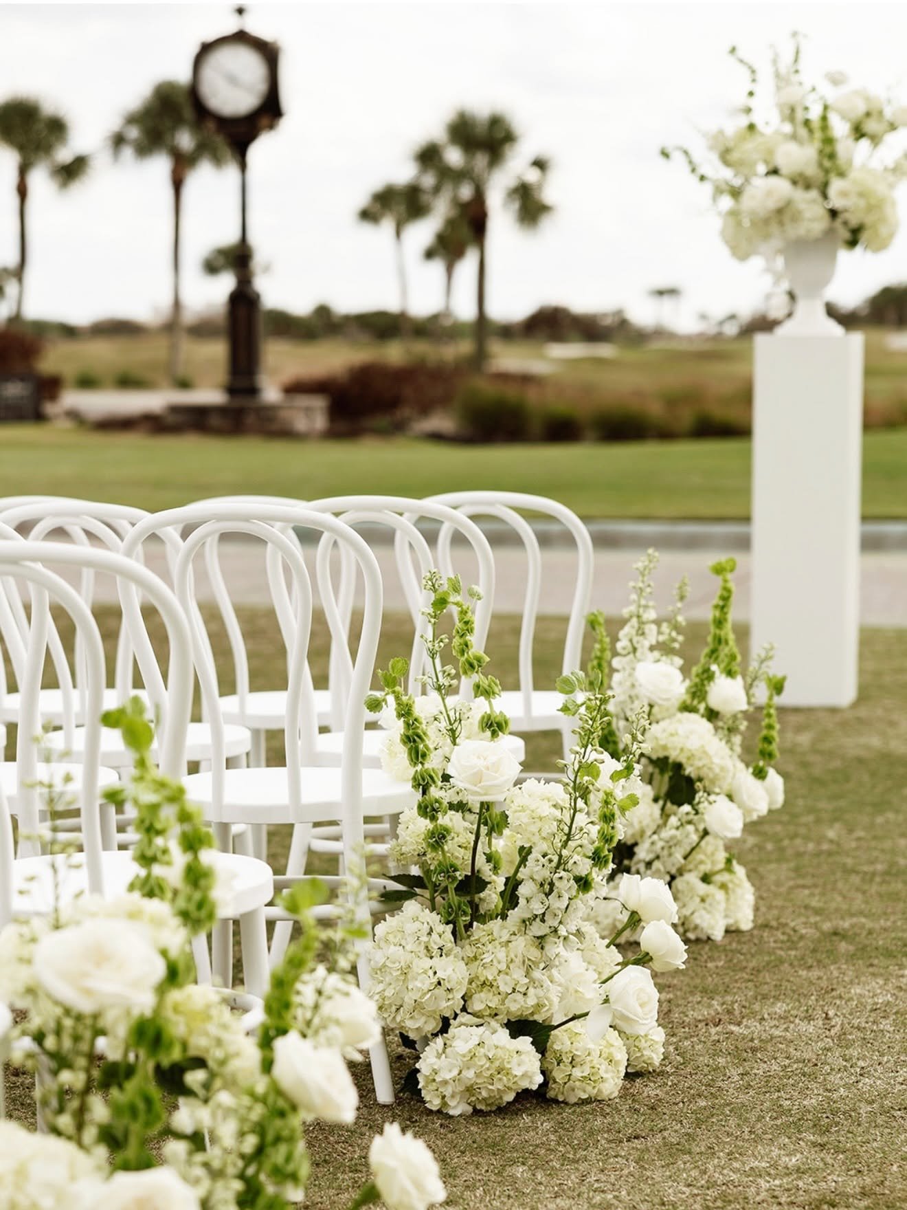 Outdoor wedding setup with white chairs decorated with white and green floral arrangements, a flowered arch, and a vintage clock in the background.