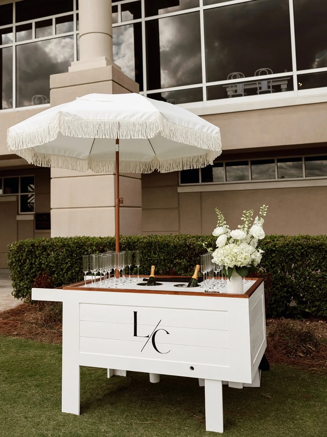 A white outdoor bar cart with bottles of champagne, surrounded by glasses and a floral arrangement, set up for a celebration, in front of a modern building. A large white umbrella provides shade.