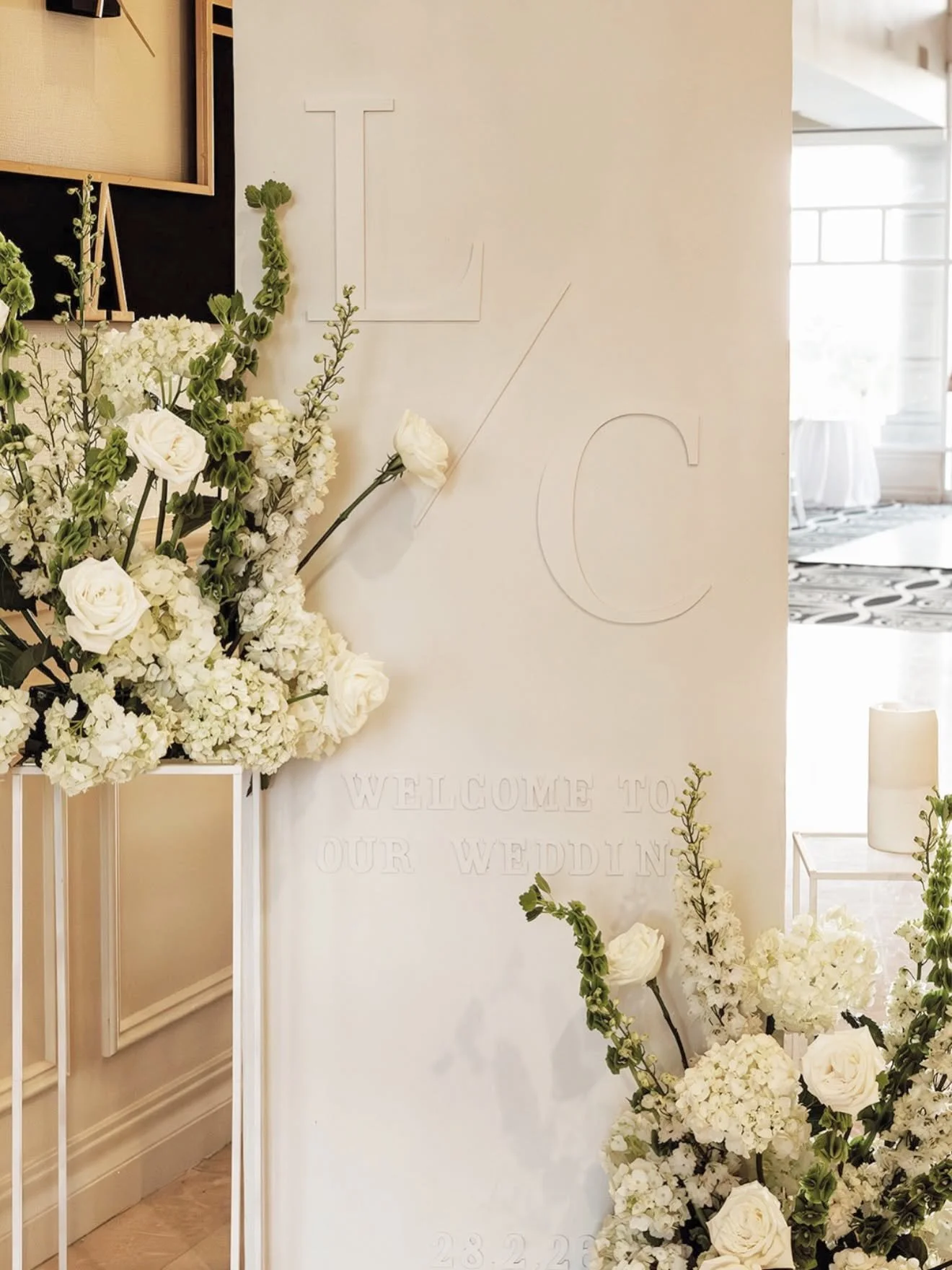 White wedding welcome sign with white flowers and greenery, featuring the word 'LOVE' outlined on the wall behind with the message 'Welcome to our wedding' and the date '28.2.23'