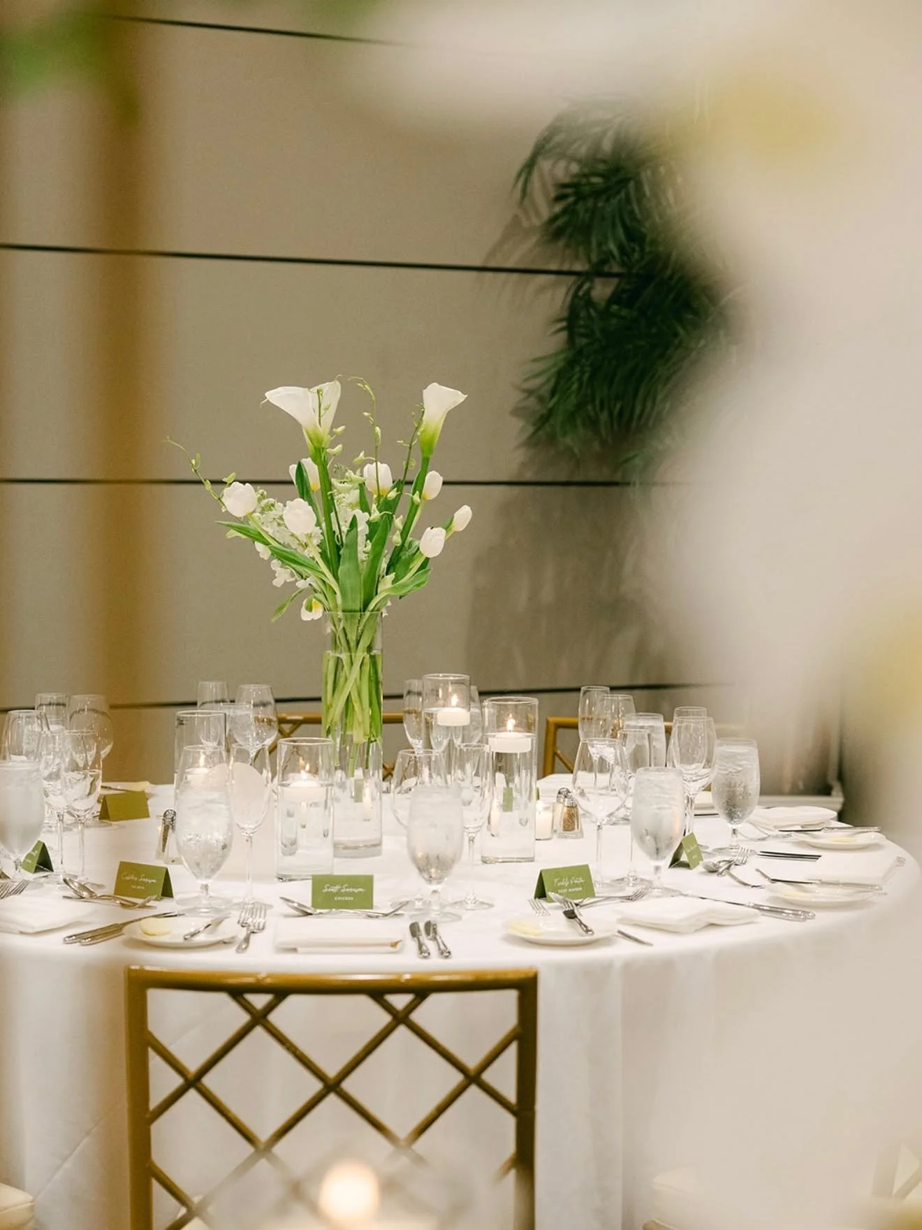 Elegant banquet table setting with a tall floral centerpiece of white lilies and greenery, surrounded by wine glasses, water glasses, and place settings in a modern decorated venue.