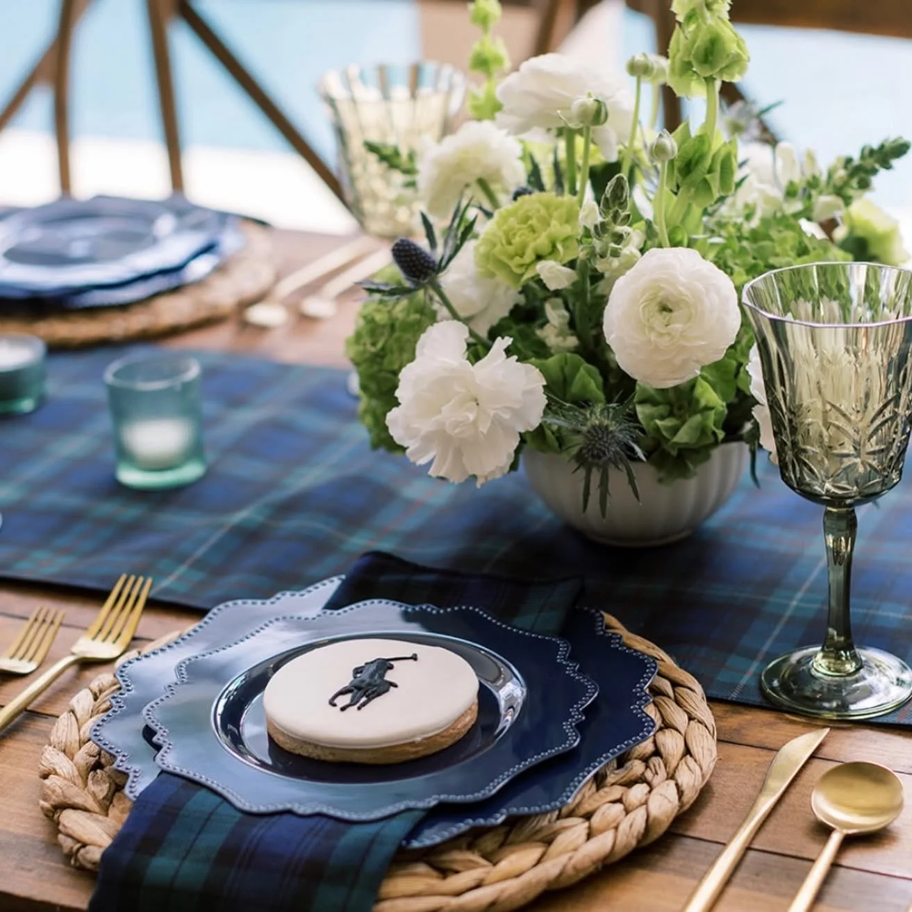 A dining table decorated with a floral centerpiece of white and green flowers, set with gold utensils, blue patterned plates, a cookie with a football decoration, a blue plaid table runner, and a glass of wine. Candles and a woven placemat are also v