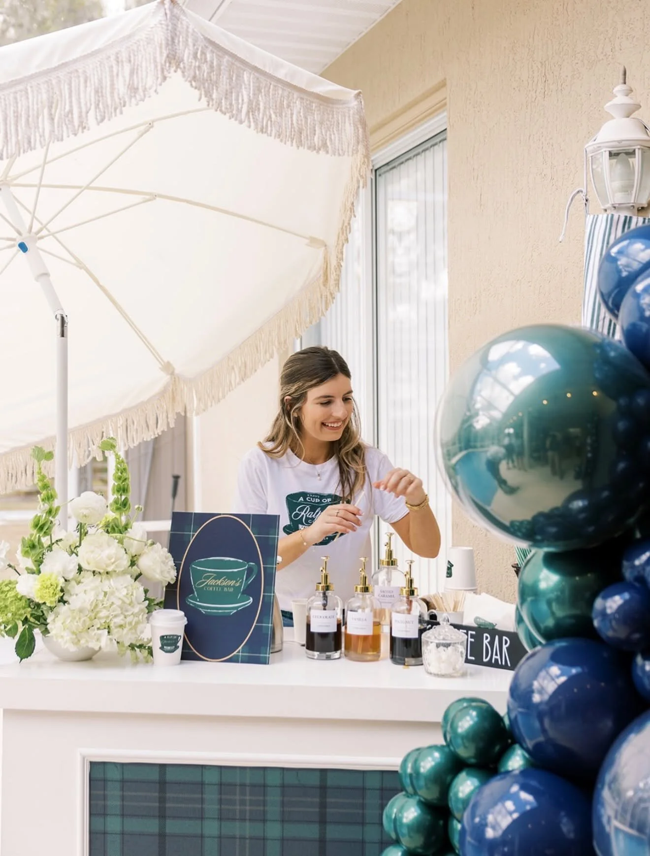 A woman smiling behind a booth selling coffee. The booth is decorated with a bouquet of white flowers, a blue and green plaid sign with a coffee cup logo, and a balloon display in shades of blue and green.