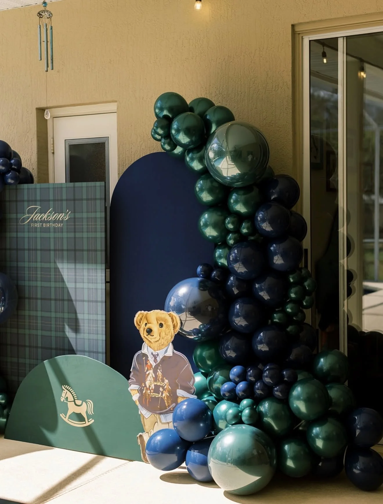 Party decoration with a balloon arrangement of green, dark blue, and light blue balloons next to a cardboard cutout of a dog dressed in a hunter's outfit, a wooden rocking horse emblem, and a backdrop with a plaid pattern and the text "Jackson's Firs