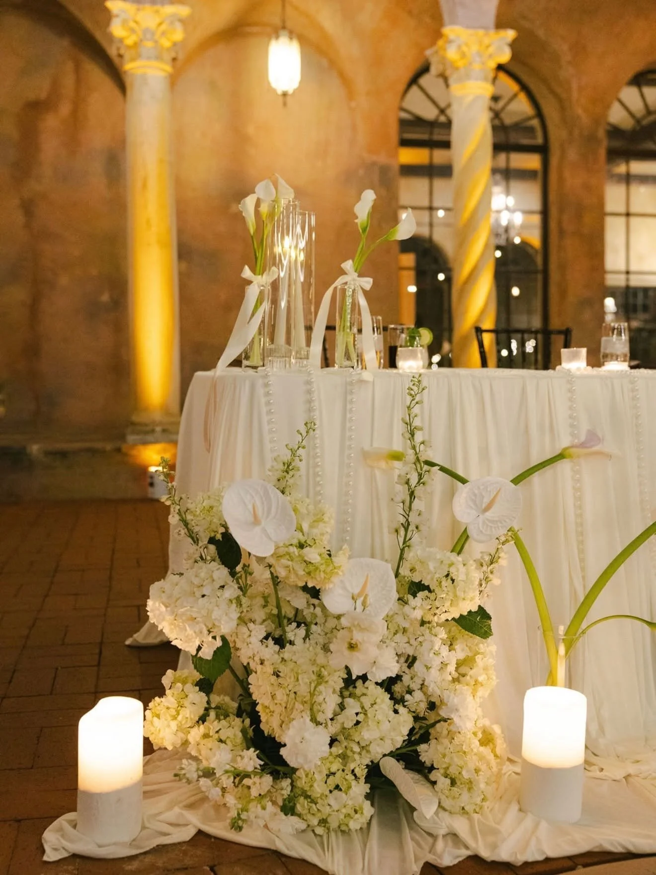 Elegant wedding or event table decorated with white flowers, candles, and glass vases in a warm, classical venue with brick walls and arched windows.
