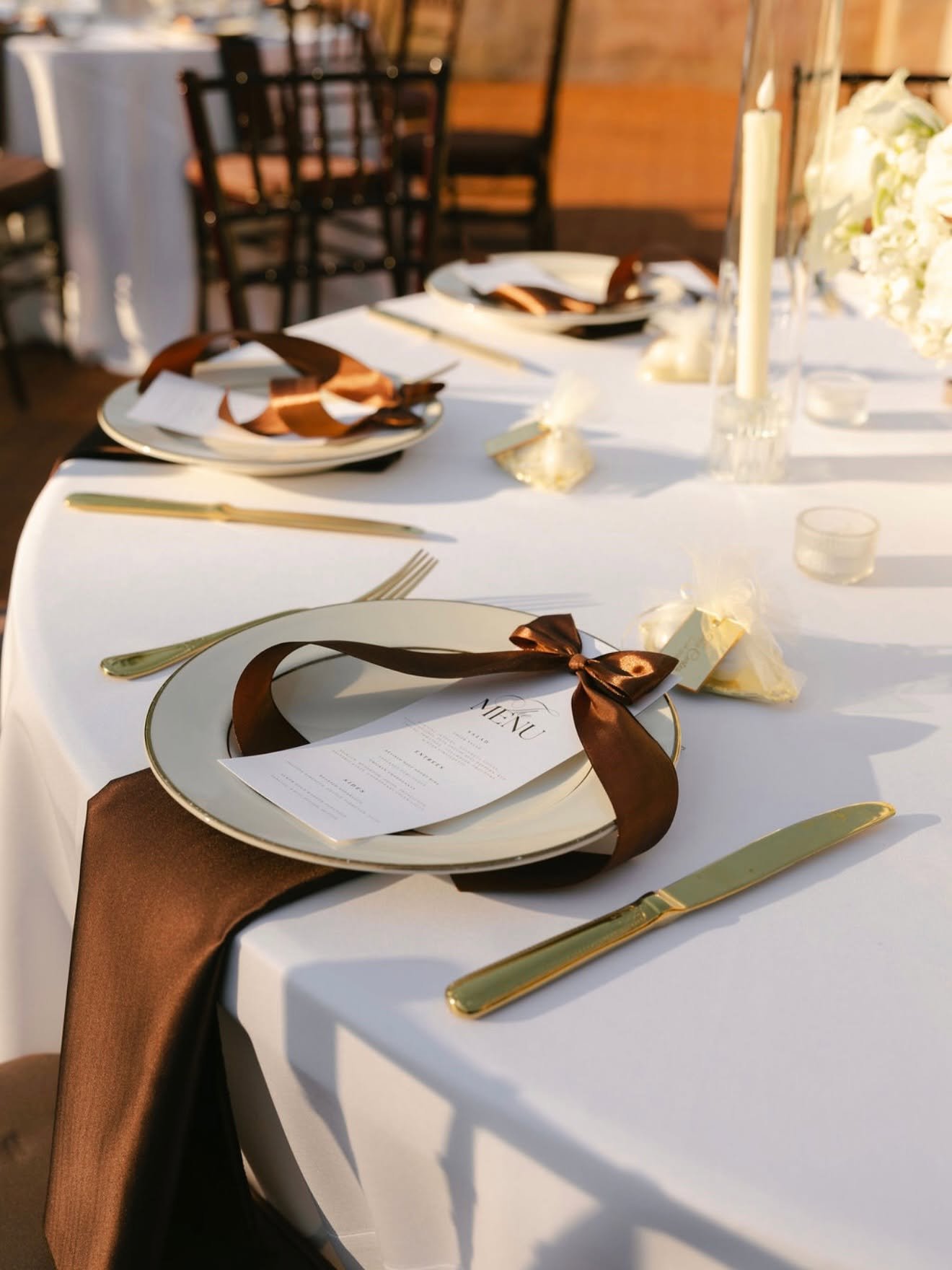 Elegant table setting at a formal event with white tablecloths, gold utensils, and brown satin ribbons tied around white plates with menus inside.