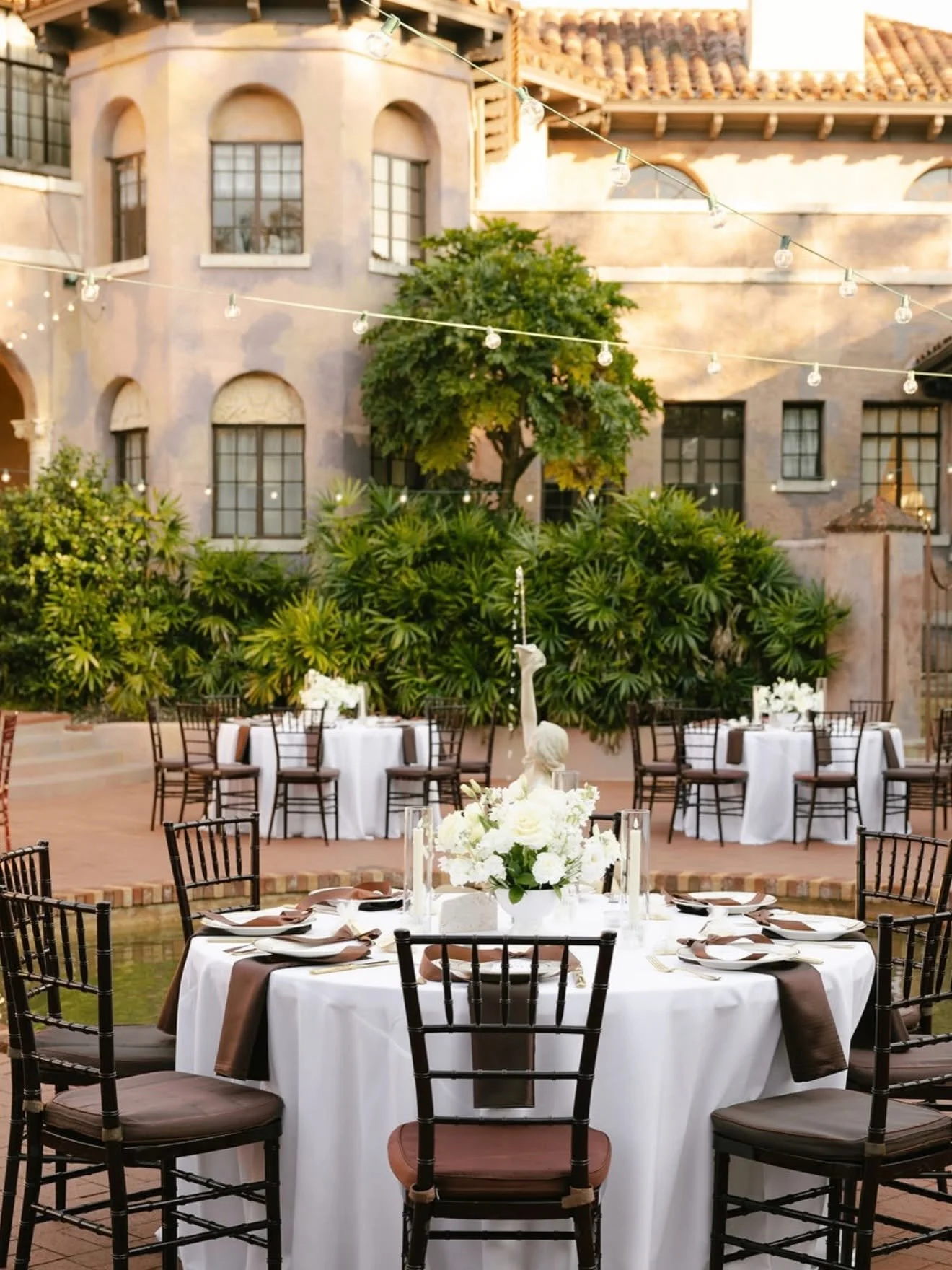 Outdoor banquet setup with round tables draped in white tablecloths, decorated with white floral centerpieces and dark wooden chairs, surrounded by lush green plants and trees, with a European-style building in the background decorated with string li