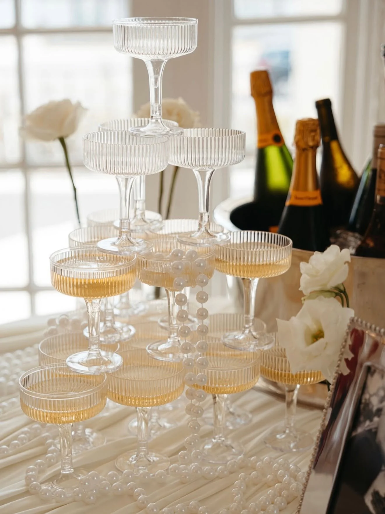 A tiered display of empty champagne glasses with a champagne bottle in an ice bucket in the background. White flowers and pearl decorations are also visible.