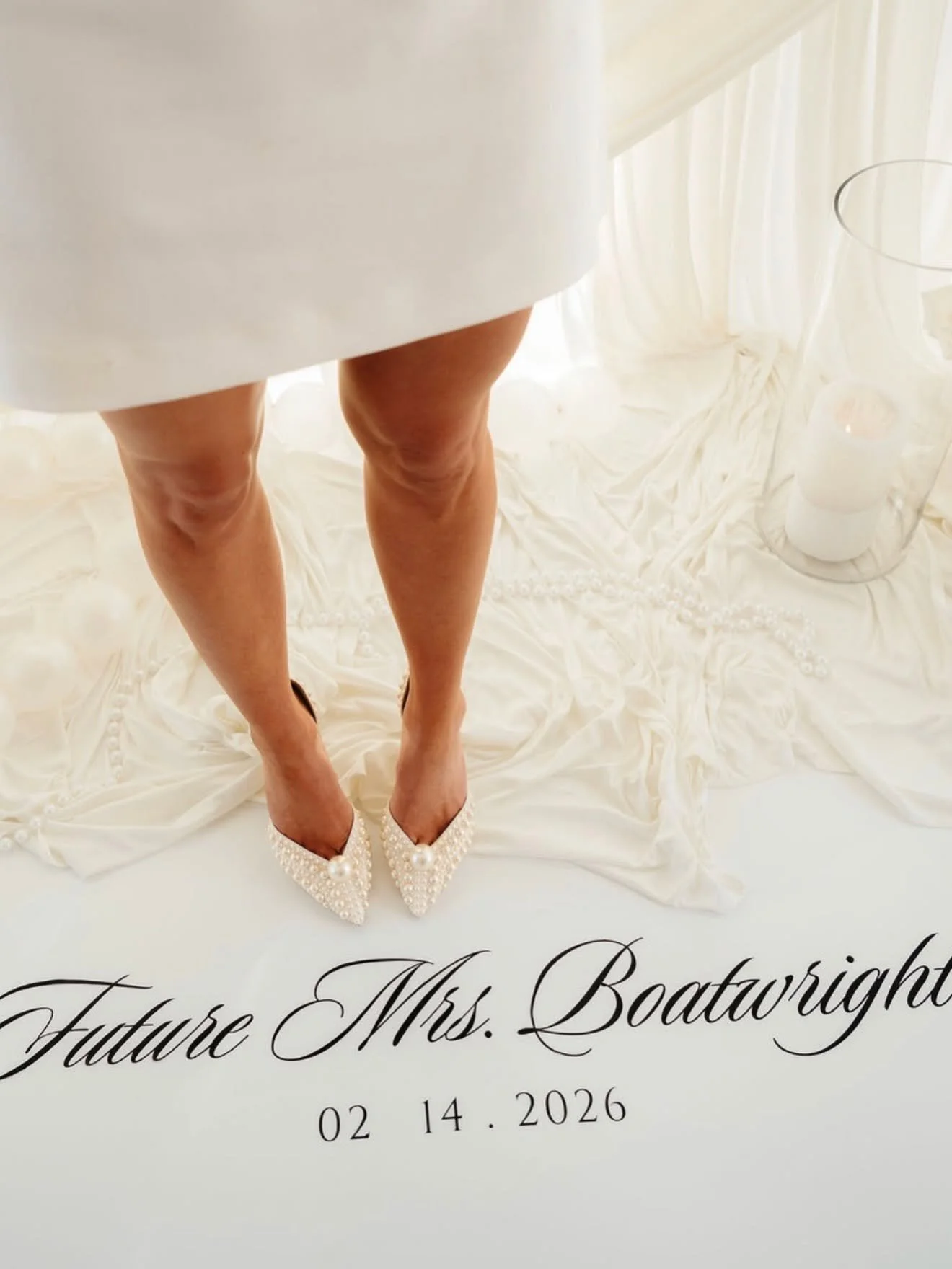 A pair of legs in pearl-embellished high heels on a white silk fabric with black cursive text reading "Future Mrs. Boatwright" and a date of February 14, 2026, with white candles and a glass on a white surface in the background.