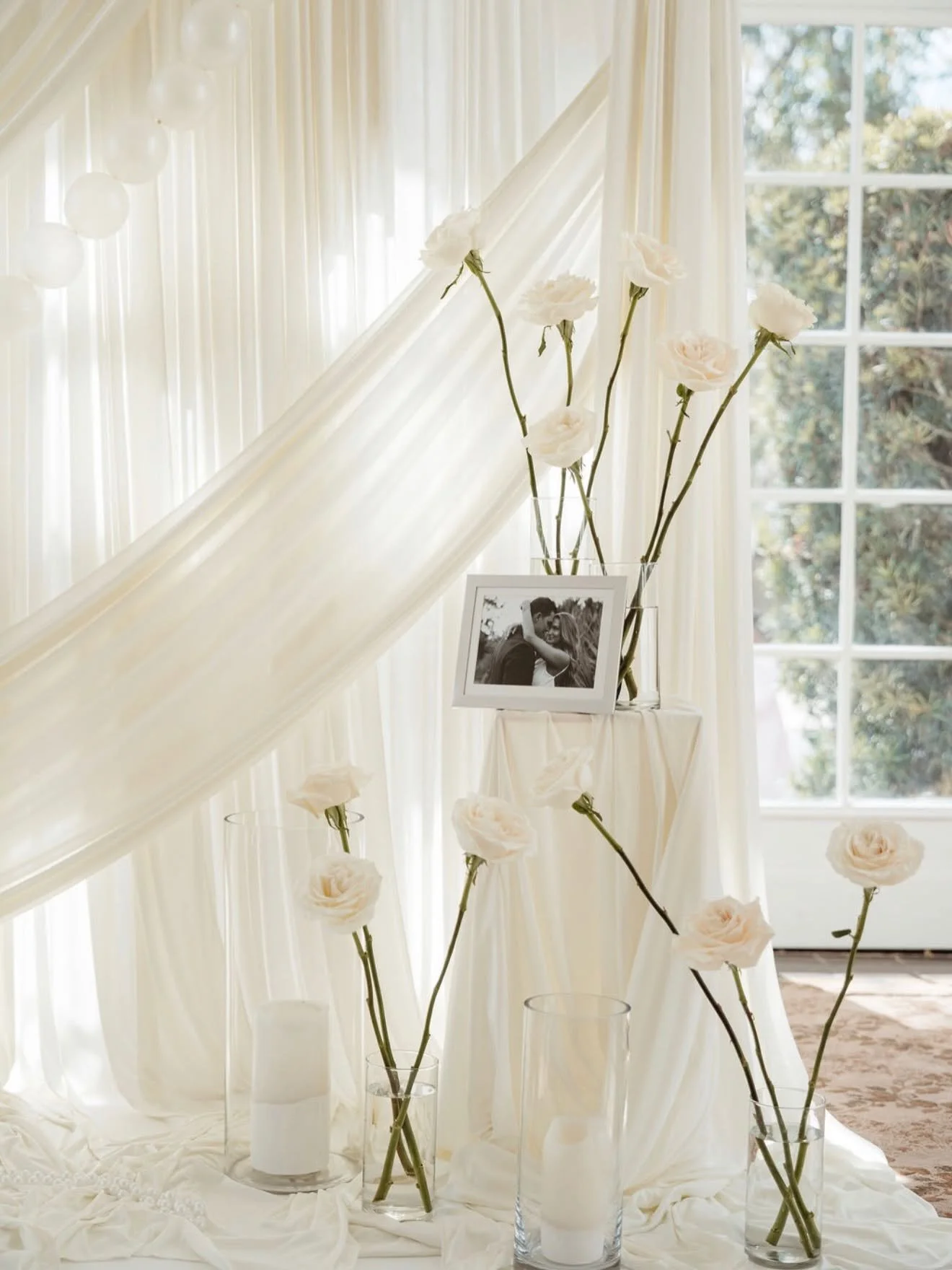 Decorative scene with white roses in tall glass vases, a black and white photograph of a couple, and white curtains with natural light coming through a window.