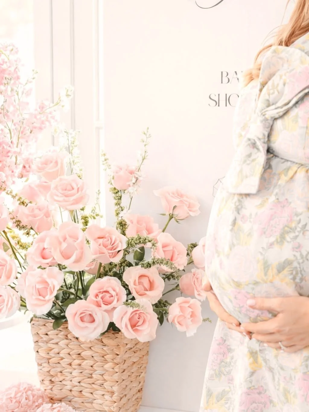 A woman with blonde hair holding her pregnant belly stands beside a basket of pink roses and other flowers inside a bright, softly lit space.