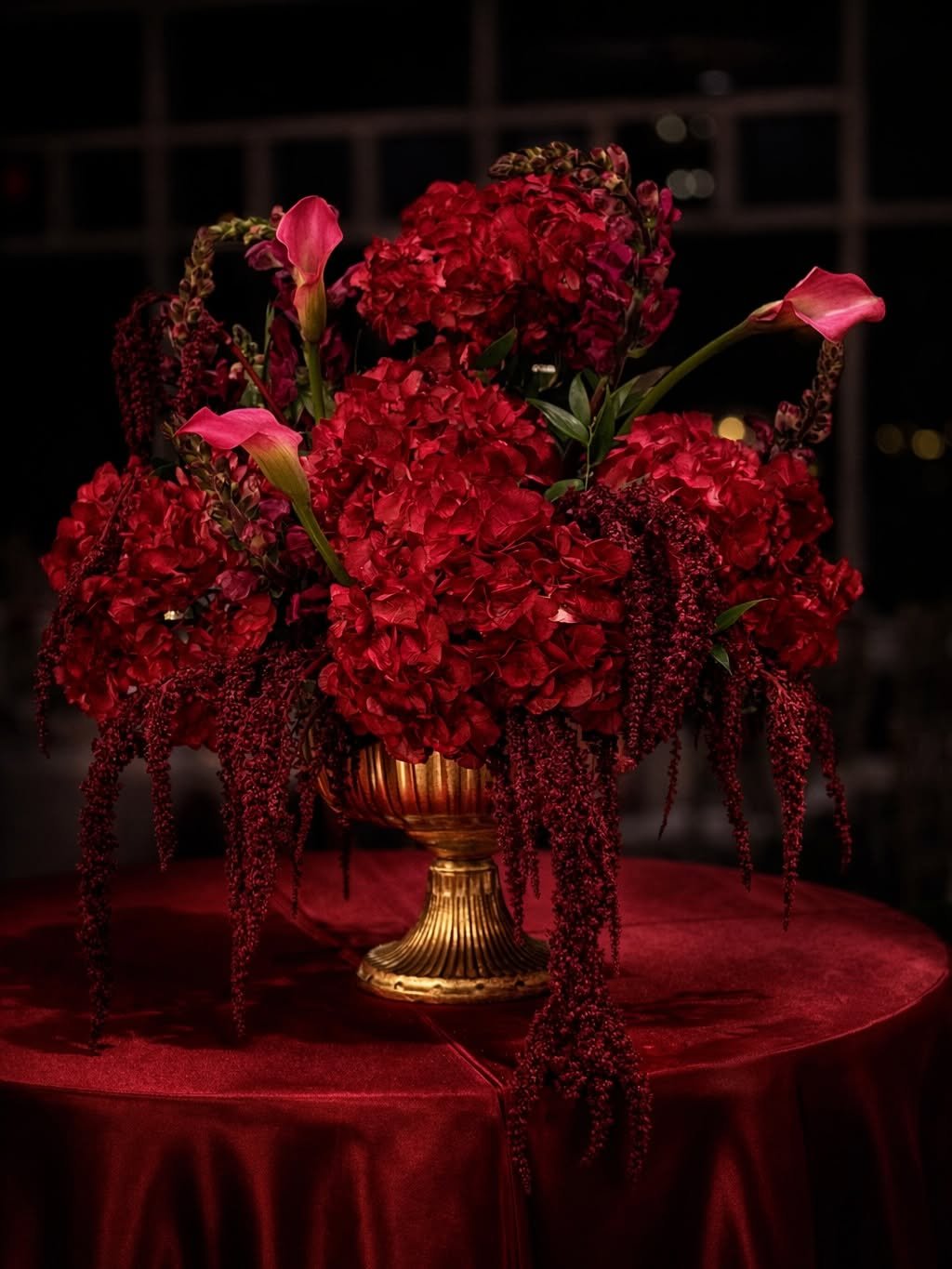 Red flower arrangement with hydrangeas and calla lilies in a gold vase on a red tablecloth, dark background.