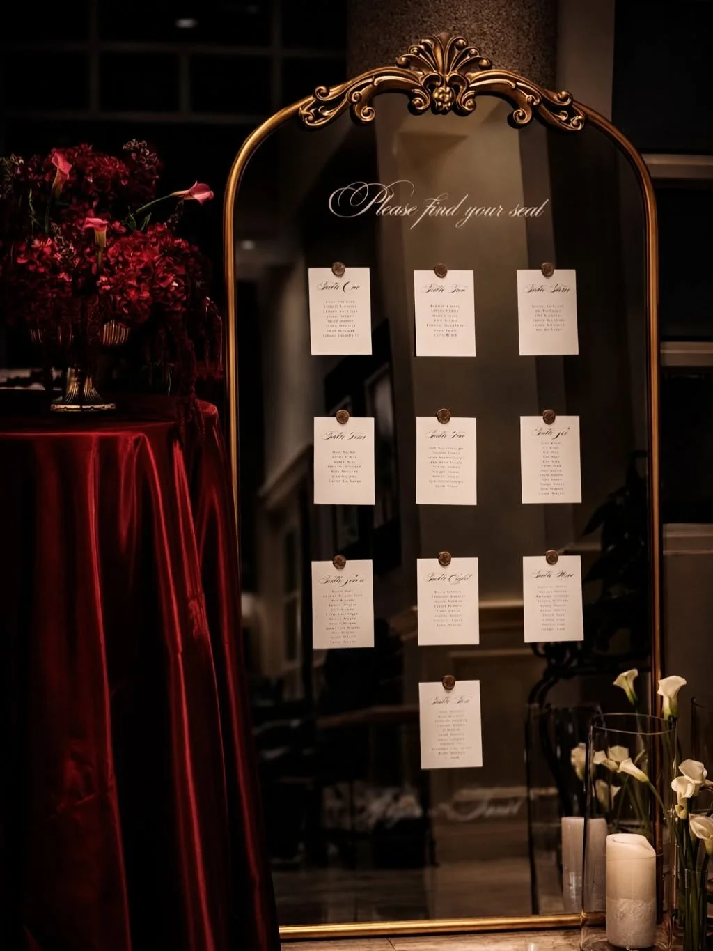A decorative mirror with a gold frame and a sign that says "Please find your seat" at a wedding or event. Below the sign are several cards with seating arrangements, pinned to the mirror. To the left, there is a bouquet of deep red flowers on a table