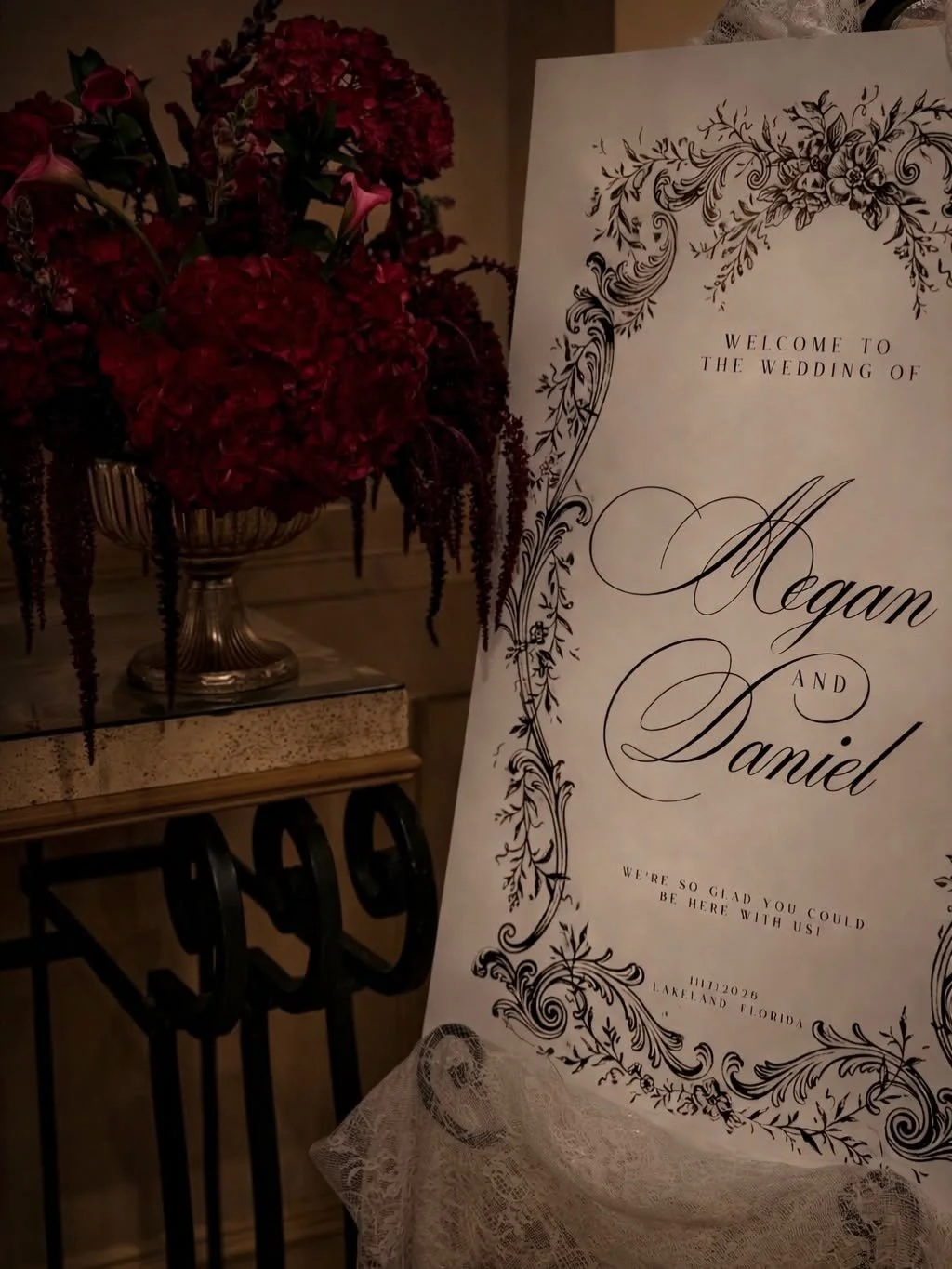 A wedding welcome sign with elegant black floral designs and the names Megan and Daniel, along with a flower arrangement of deep red hydrangeas and pink calla lilies in a silver vase.