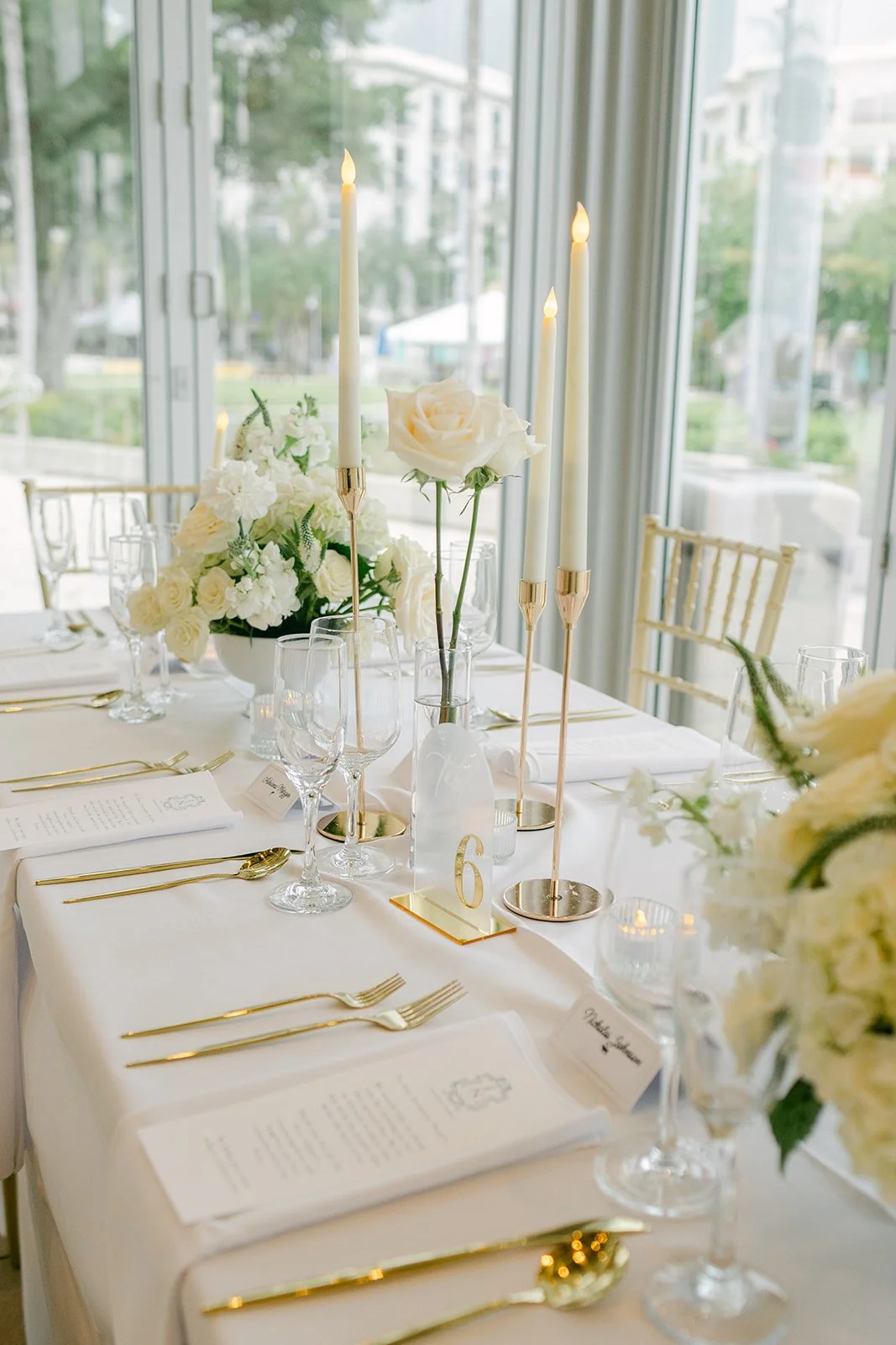 Elegant table setup with white flowers, tall candles, and gold utensils in a bright room.