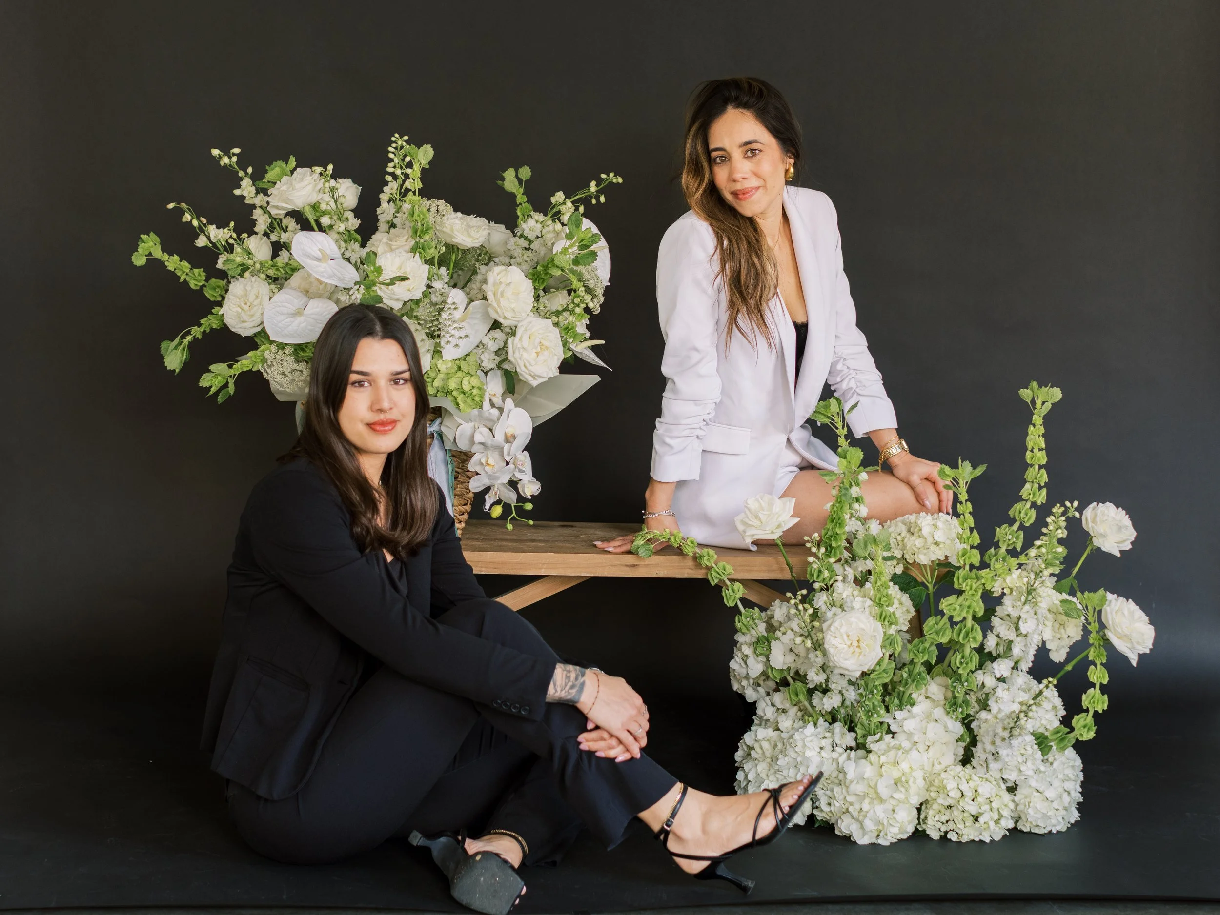 Two women with dark hair and fair skin posing with white flowers against a dark background. One woman is sitting on the floor wearing a black blazer and black pants, and the other is standing with one knee on a wooden table, wearing a white blazer and black lace top. The scene is decorated with large white floral arrangements and greenery.