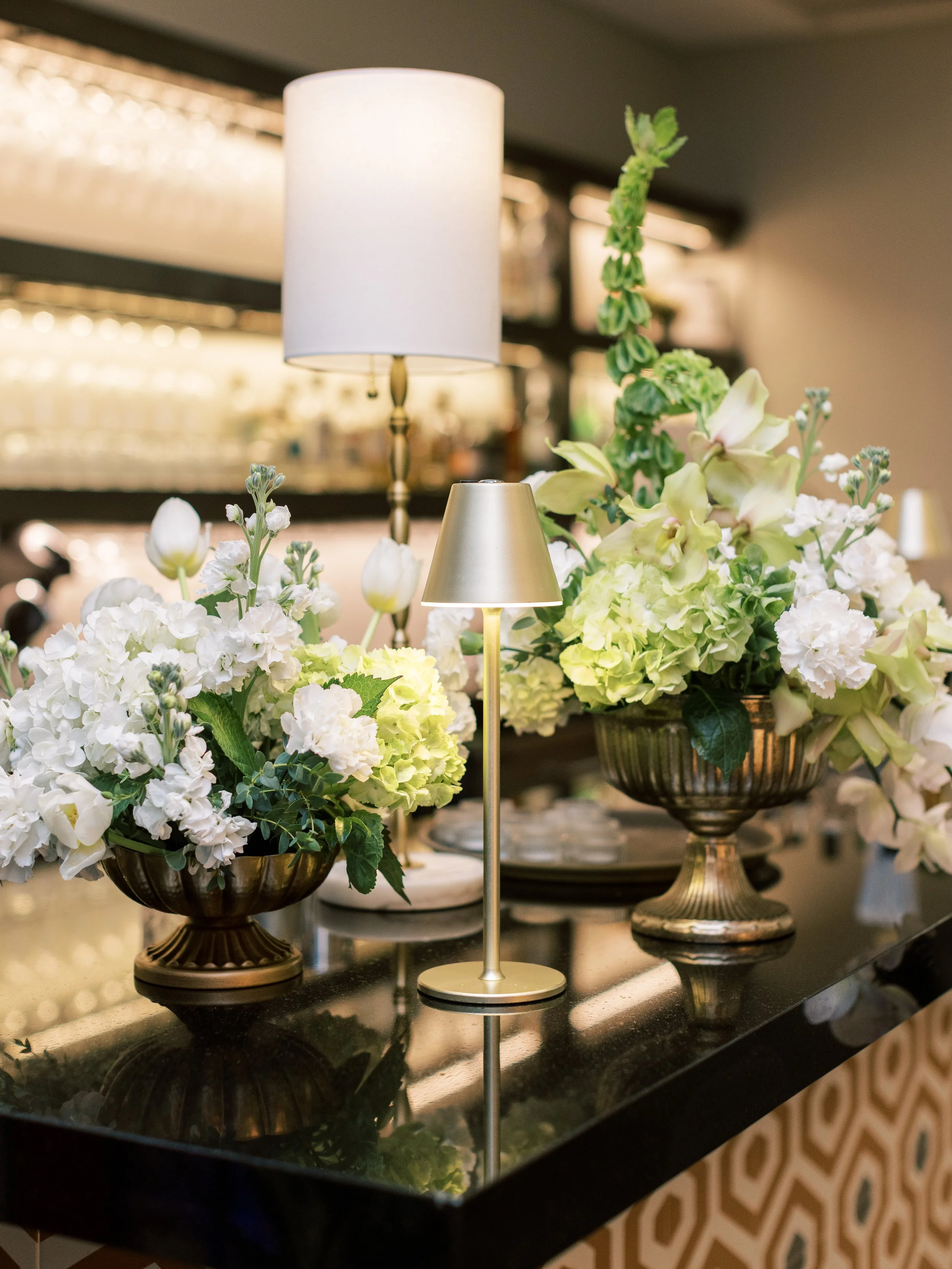 A table display with white flowers in decorative bowls, a small brass-colored lamp, and a tall white lampshade on a gold stand, set on a dark polished surface.