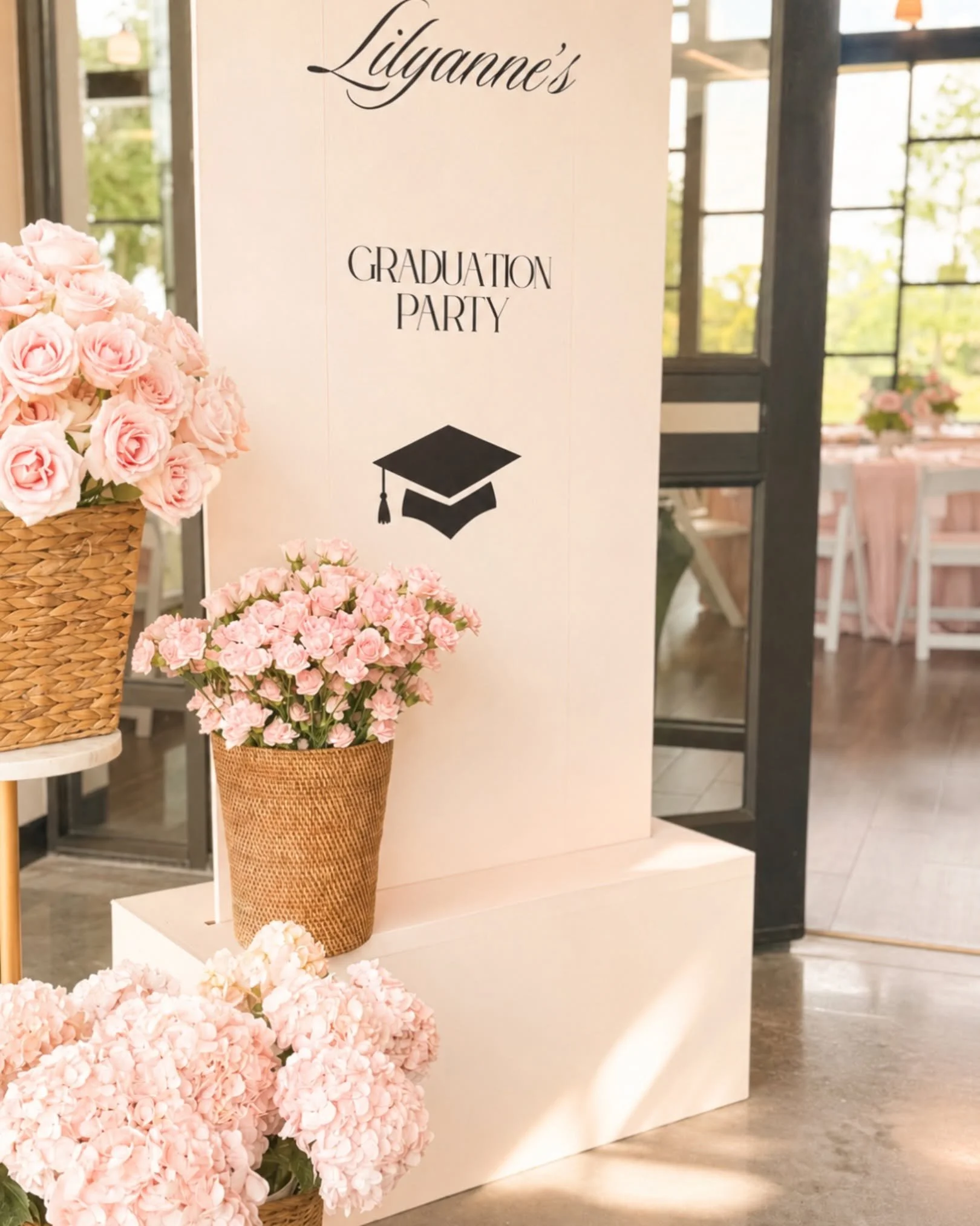 A wedding or graduation party display with pink flowers in baskets and a white sign that reads 'Lilyanne's Graduation Party' with a graduation cap graphic.