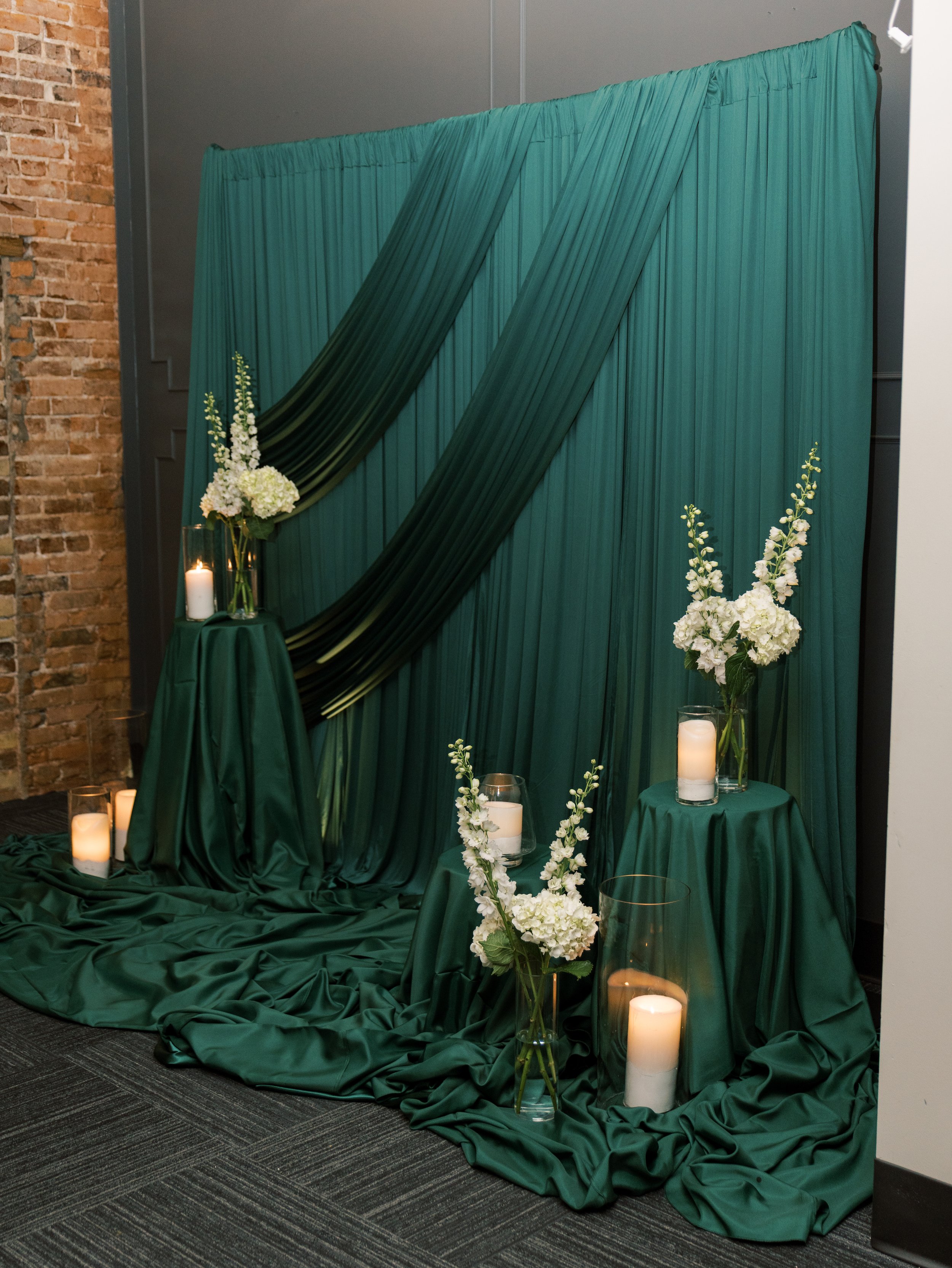 Decorative setup with green fabric backdrop, white flowers in vases, and candles on tables and floor, likely for an event or celebration.
