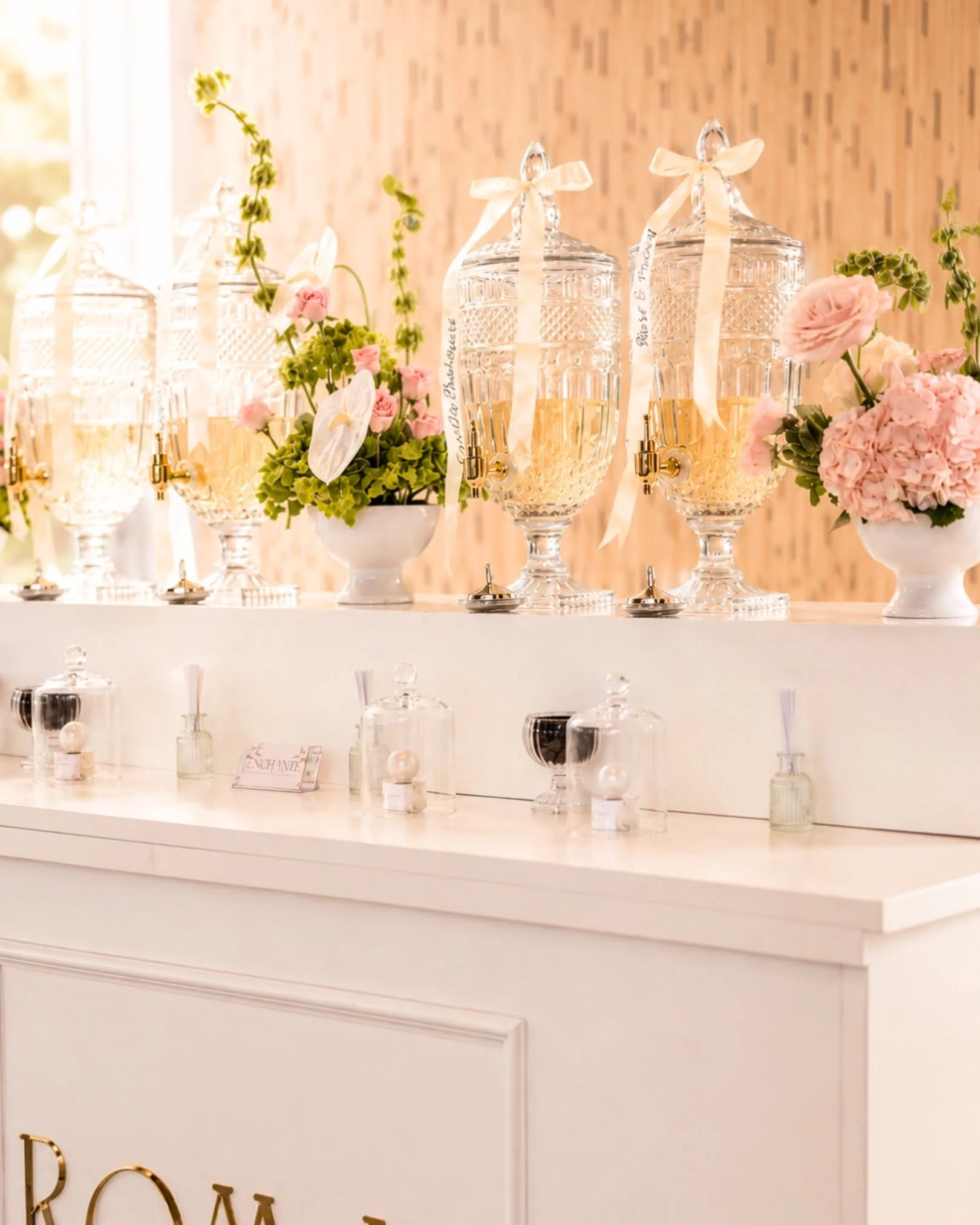 Glass beverage dispensers with ribbons and floral arrangements on a white table, set against a brick wall background for a celebration or party.