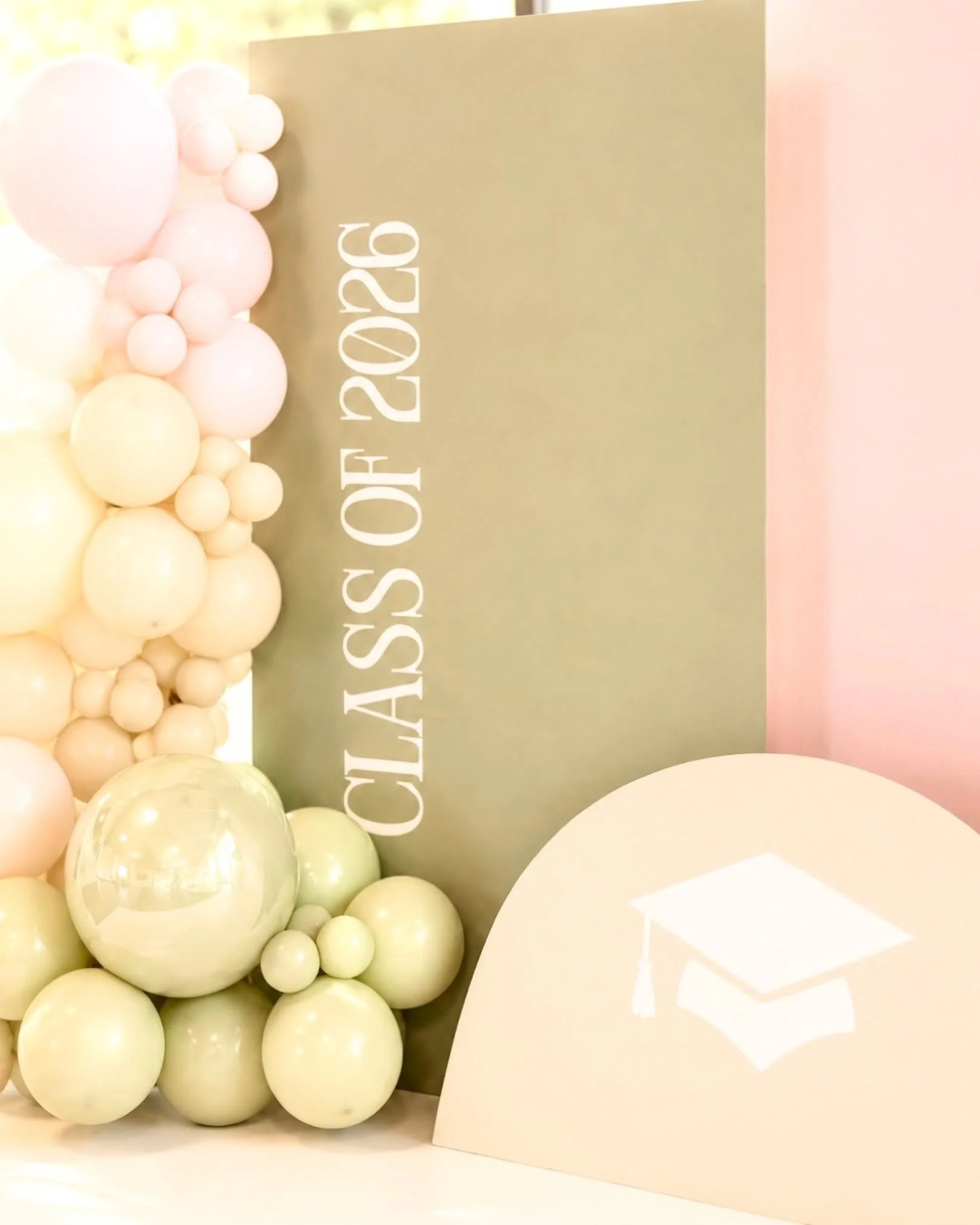 A display of white and pale yellow and green balloons and a pink backdrop with a beige sign that says 'Class of 2026'
