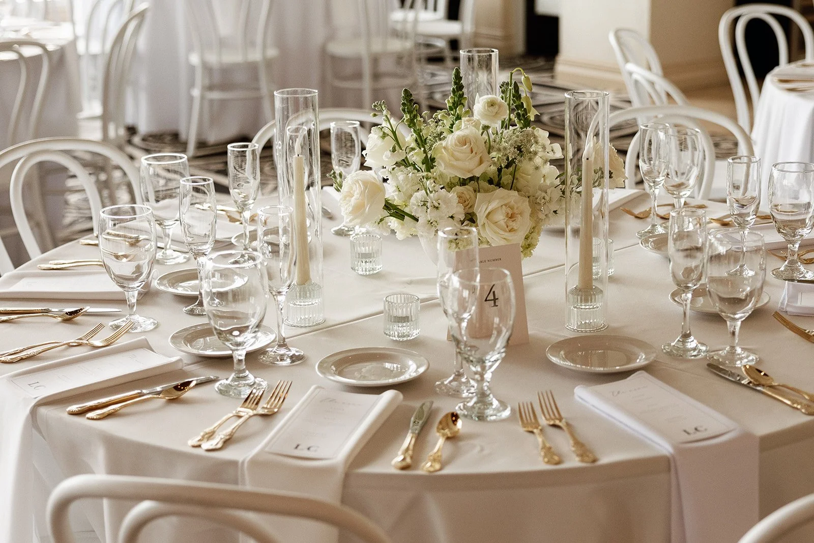 Elegant round table set for a formal event with a white tablecloth, gold cutlery, and an elaborate floral centerpiece featuring white roses and greenery, surrounded by white chairs in a well-lit room.