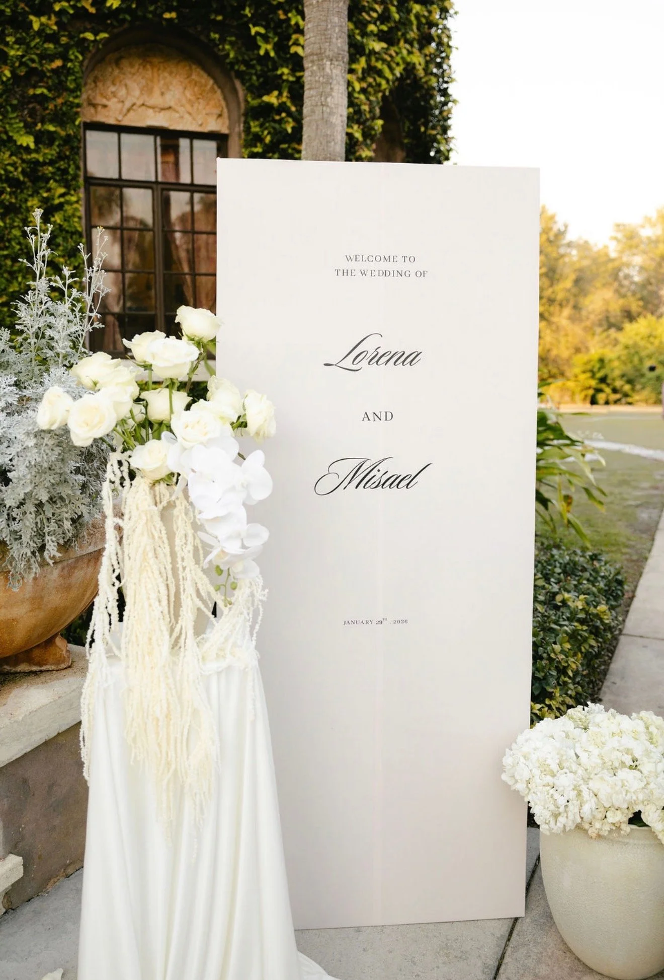 Wedding welcome sign with white floral arrangements, set outdoors with trees and a building covered in green ivy in the background.