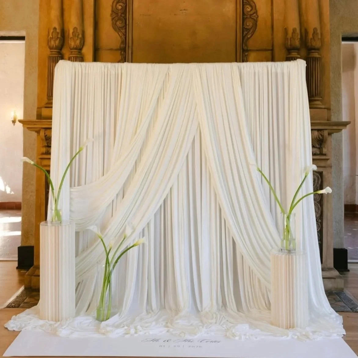 A cream-colored curtain backdrop with draped fabric, flanked by vases with white calla lilies on white pedestal stands, set in an elegant room with ornate woodwork and a fireplace.