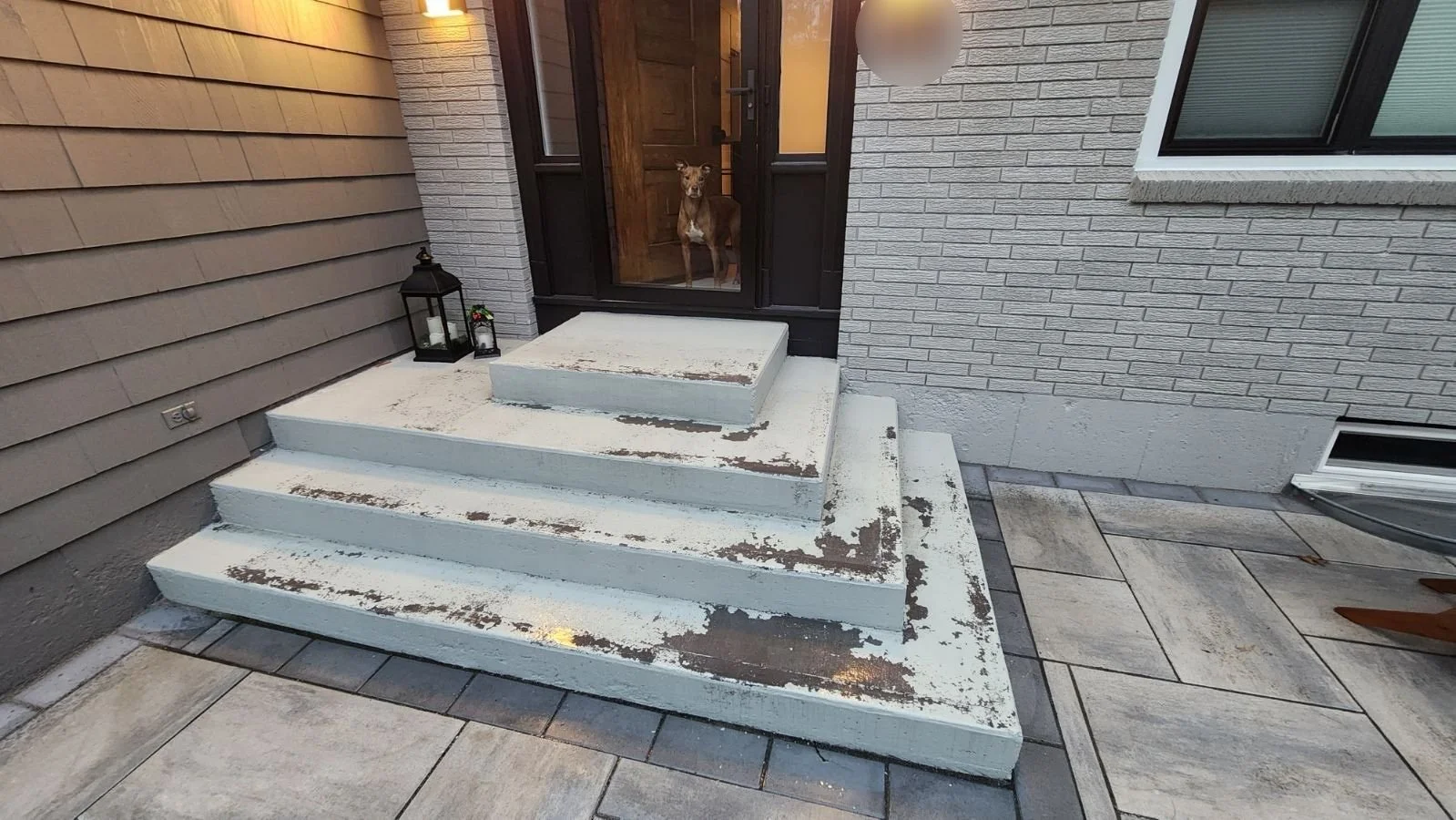 Front steps with four concrete steps leading to a glass door. A dog is visible through the screen door, sitting inside. There are two lanterns with candles on the left side of the steps. The house has beige siding and a brick wall near the door.