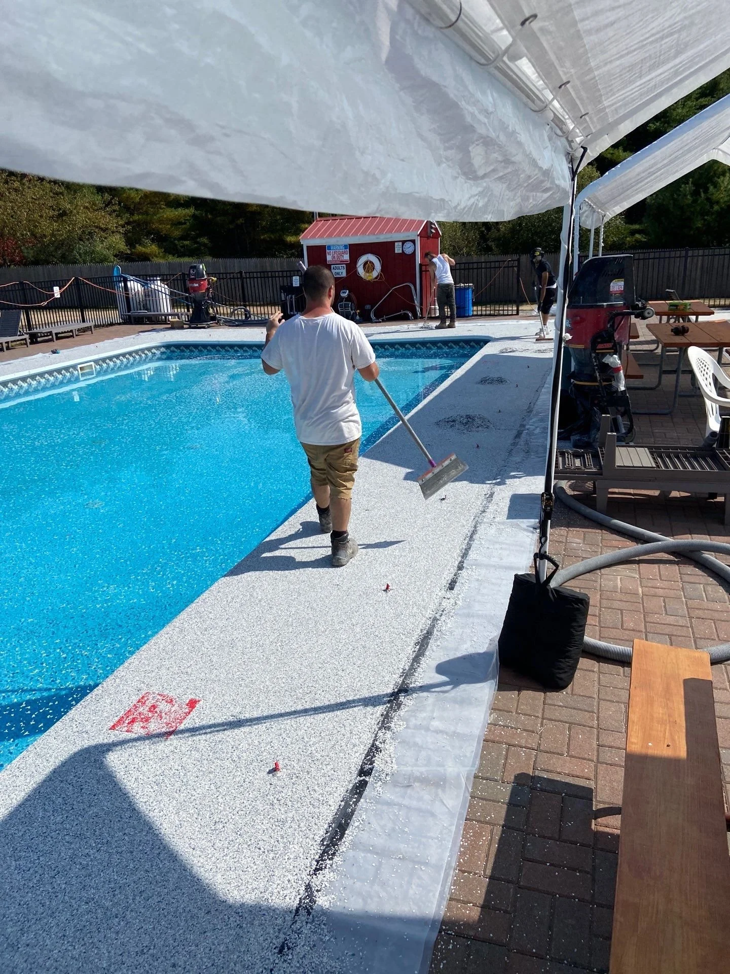 Workers cleaning the edge of an outdoor swimming pool under tents. A person in a white shirt is using a roller to apply or clean the textured surface around the pool. There are tables and chairs nearby, with equipment and fencing surrounding the pool area.