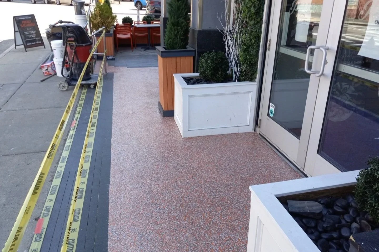 Sidewalk outside a restaurant with caution tape, potted plants, and chairs visible in the background.