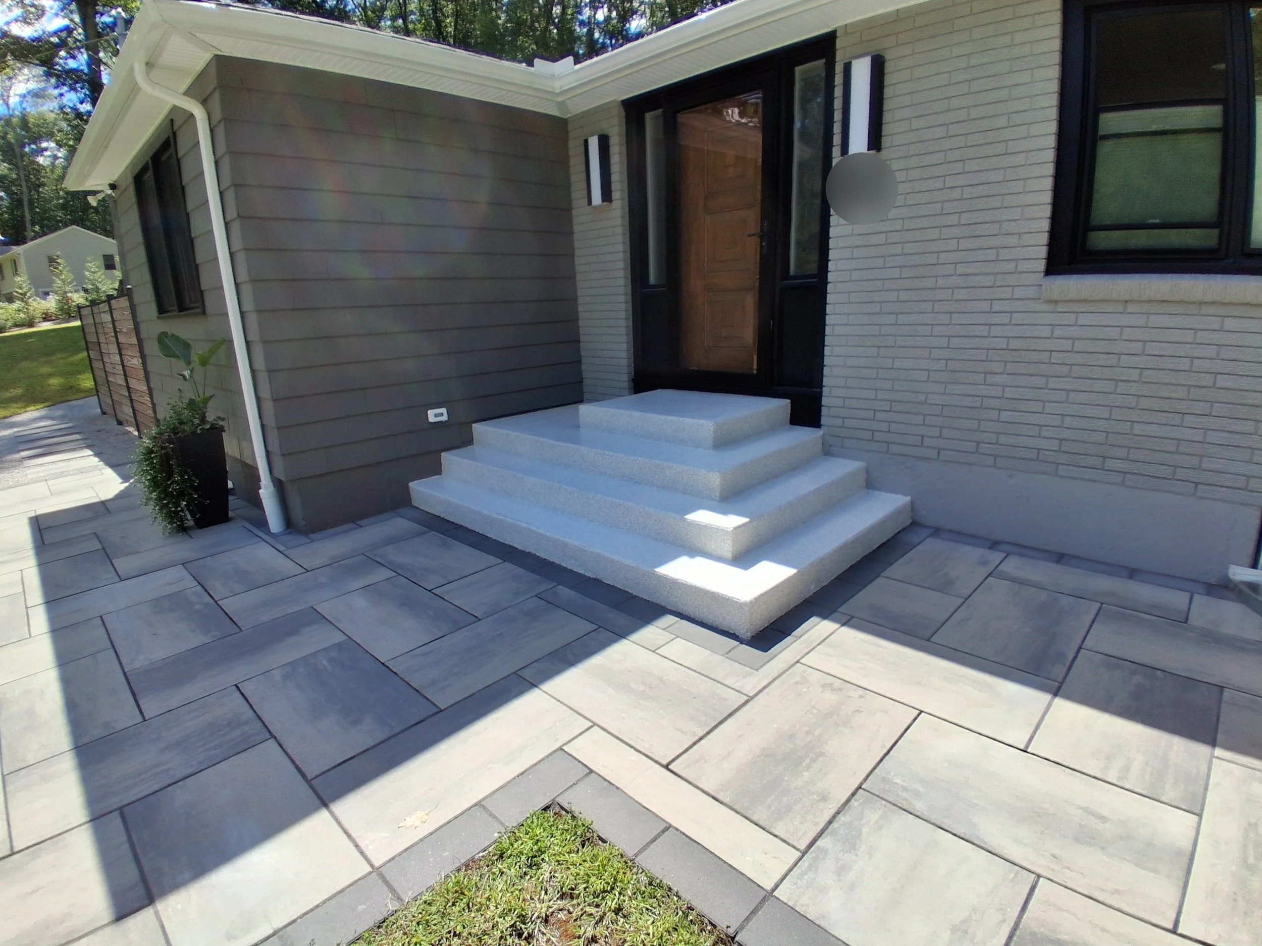 Front entrance of a house with three white steps leading to a wooden door, gray brick and beige siding walls, two vertical modern black and white lights, a potted plant, and a tiled walkway.