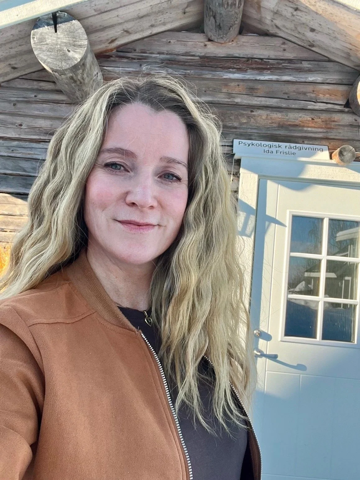Et bilde av en kvinne som smiler, med en trevegg og en dør bak henne. Det er et skilt over døren som sier 'Psykologisk rådgivning Ida Frislie'.